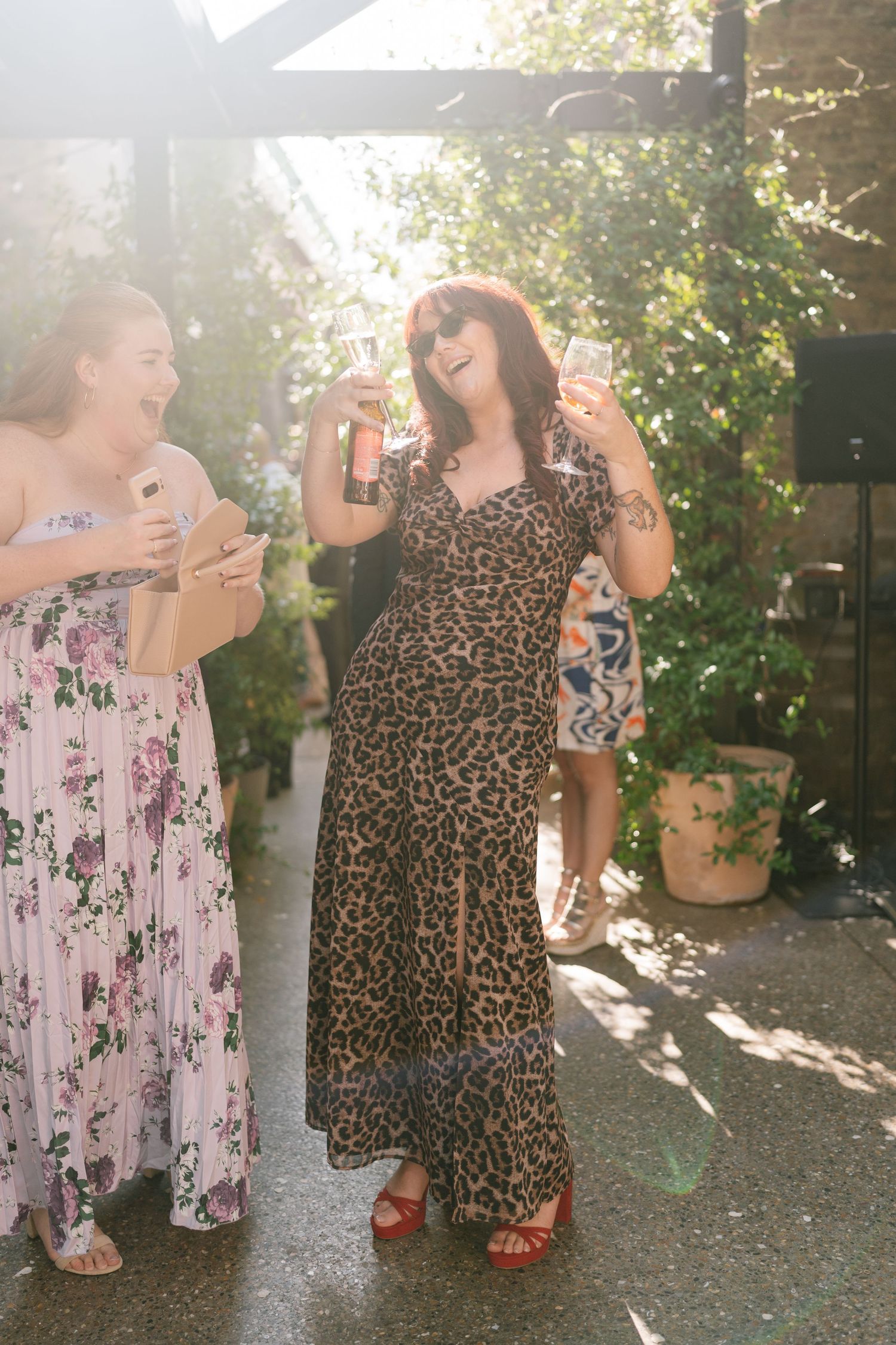 Two people in floral and leopard print dresses laugh together in a sunlit outdoor setting.