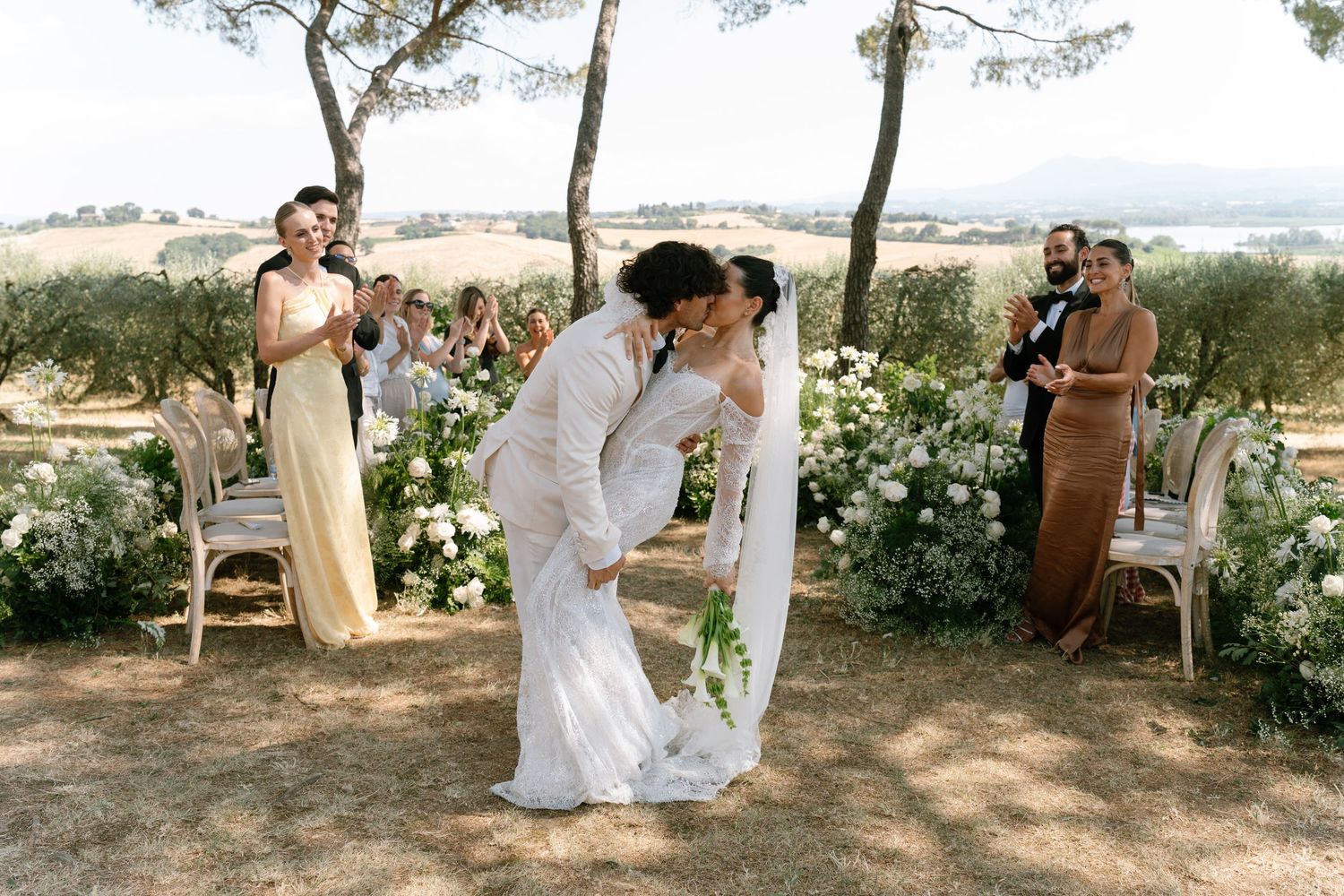 Wedding guests gather under shaded trees during an outdoor summer celebration.