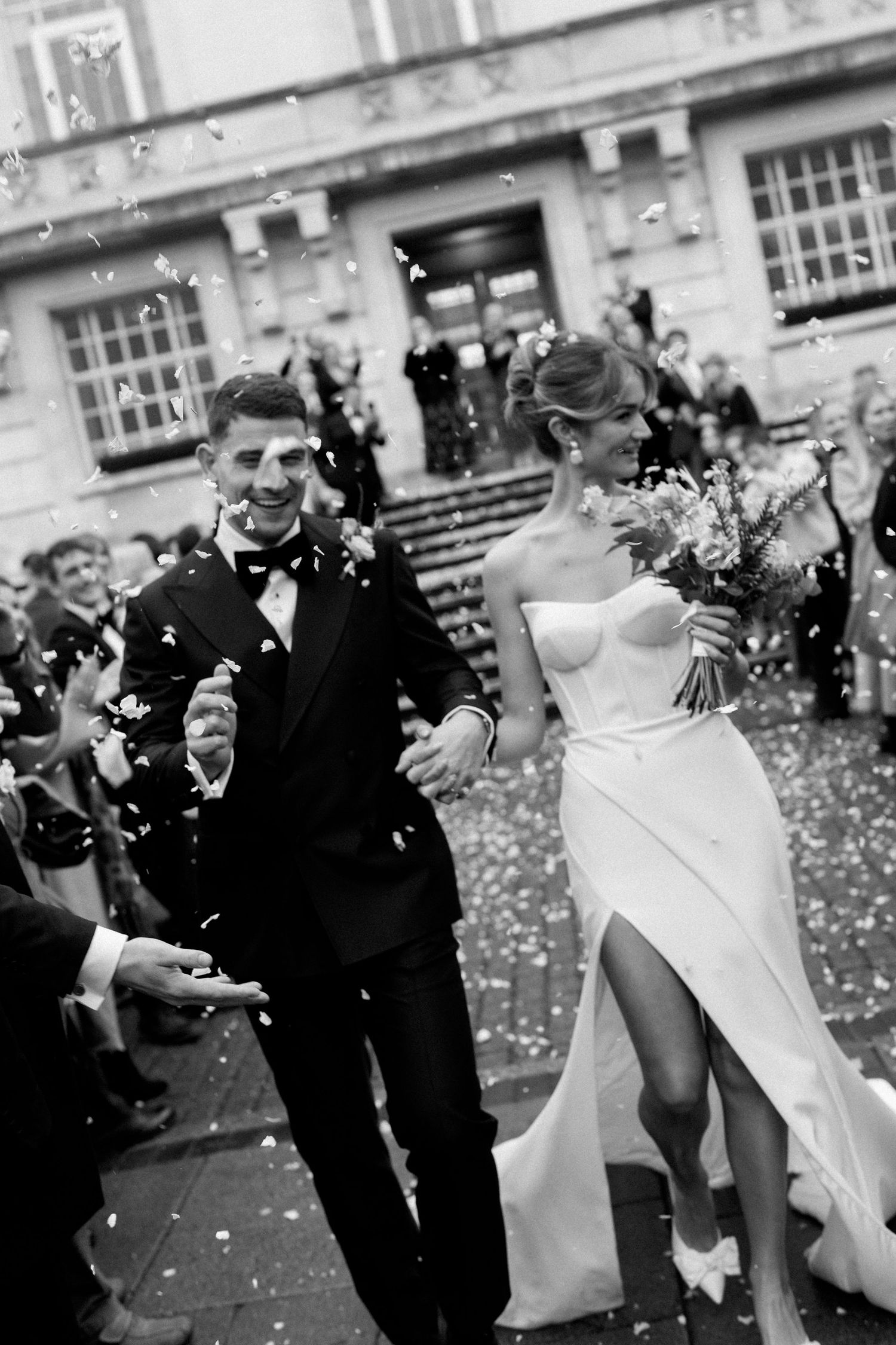 A wedding couple dances and celebrates while guests shower them with white confetti in black and white photography.