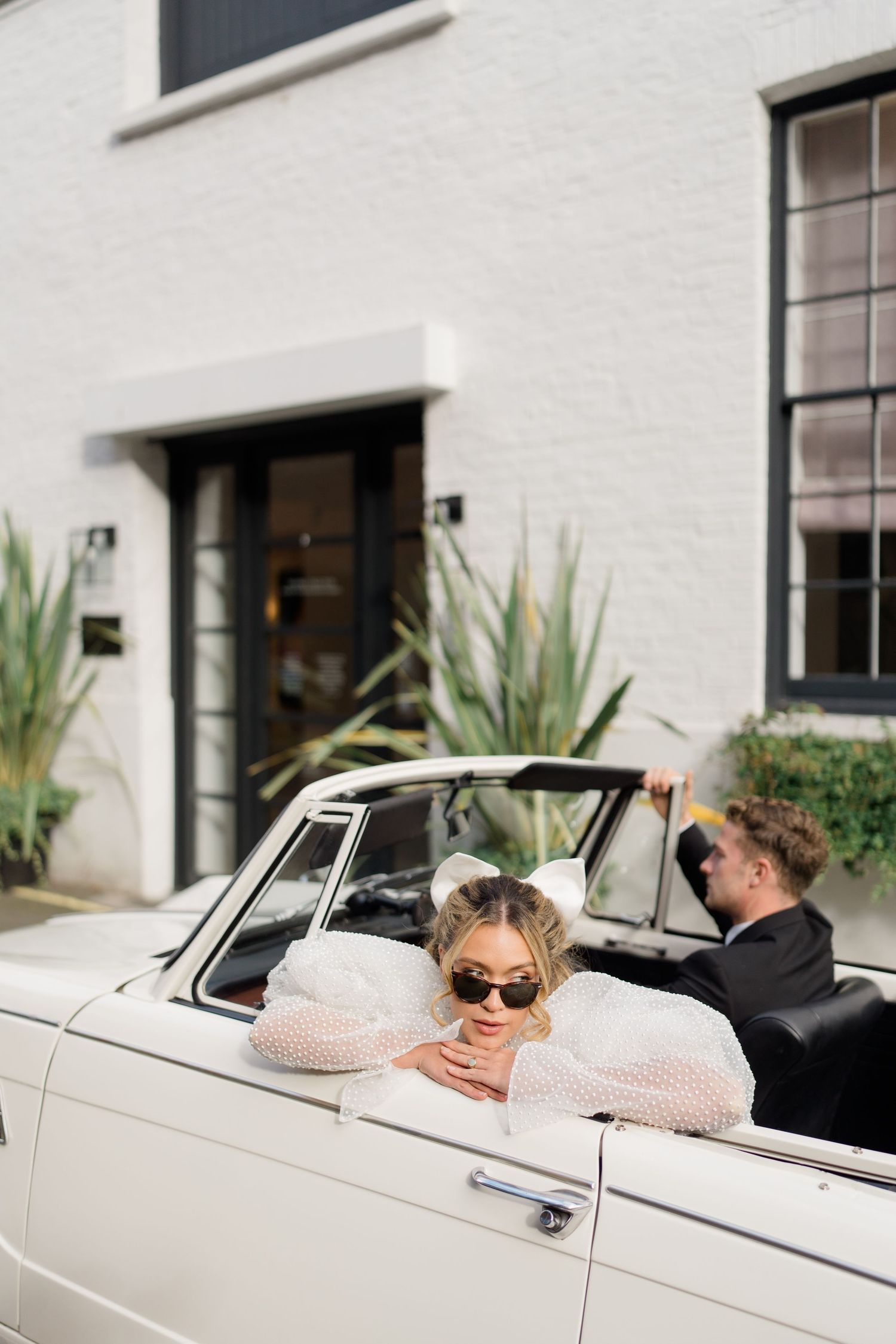 Classic white convertible car parked outside modern white building with decorative plants.