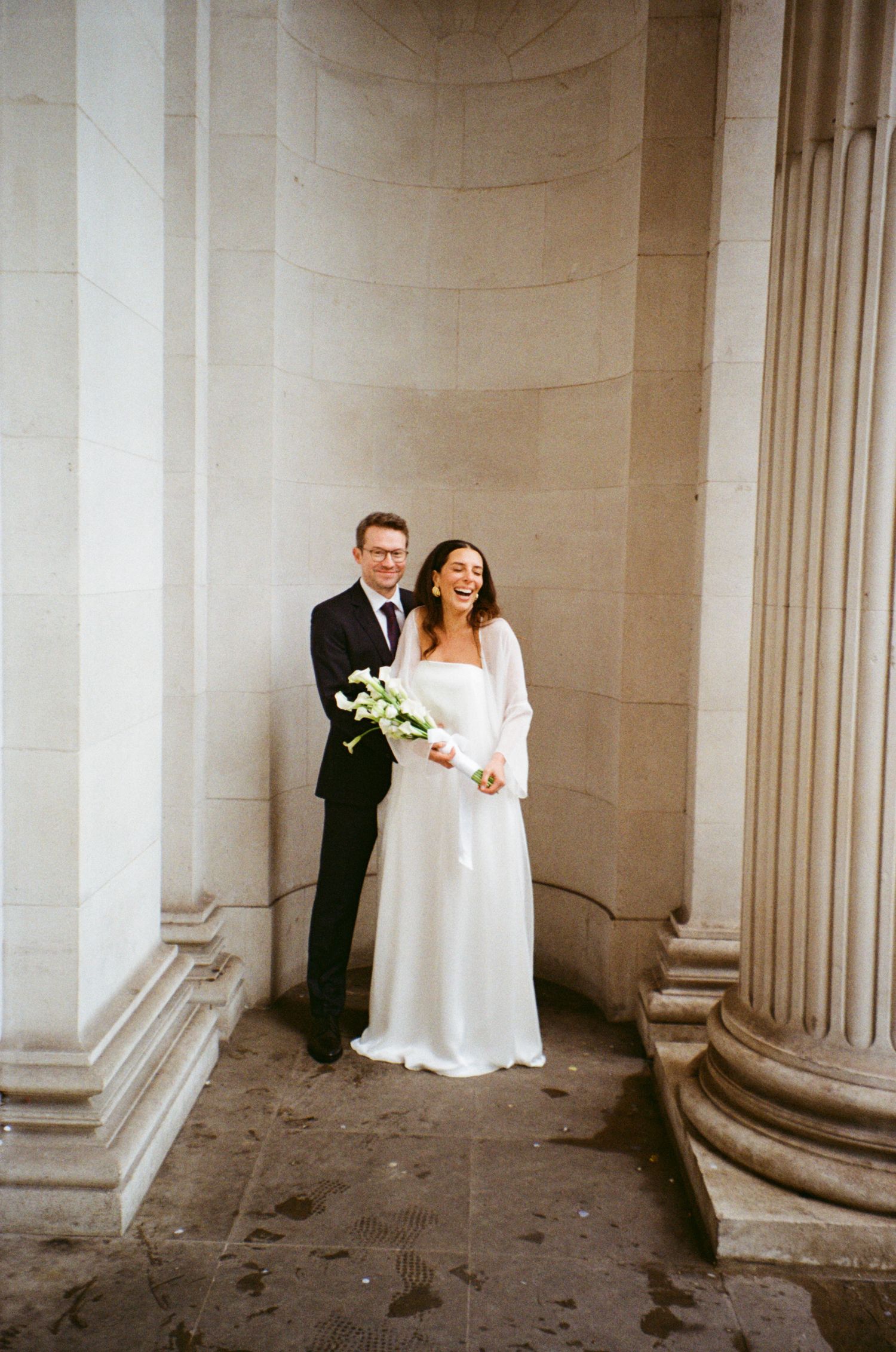 A romantic wedding photo series showing a couple posing between classical stone columns in a grand architectural setting.