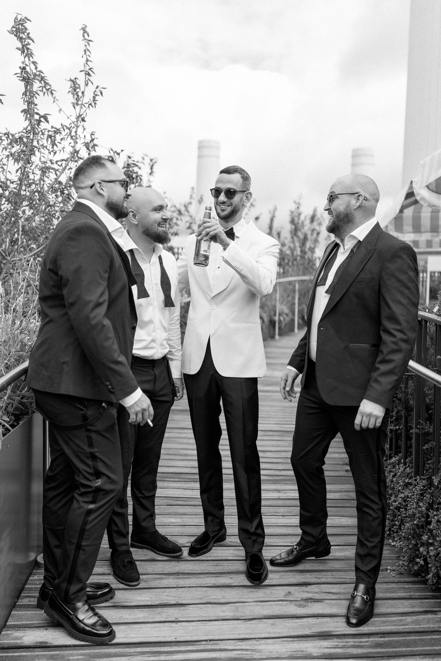 Groomsmen in formal attire share drinks and laughs on outdoor deck.
