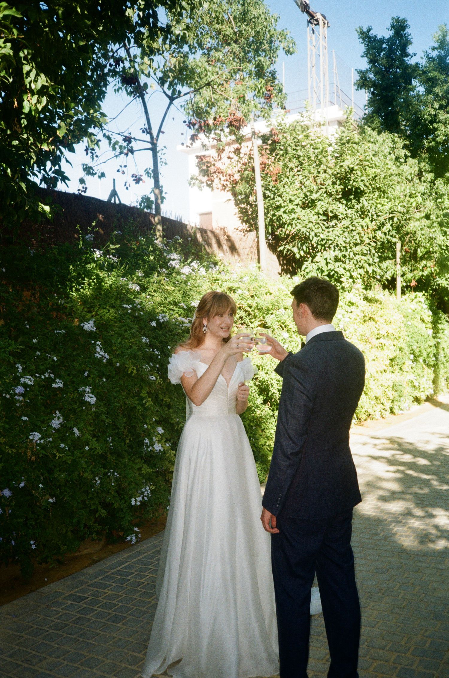 A couple in wedding attire walks together on a brick path surrounded by lush greenery on a sunny day.