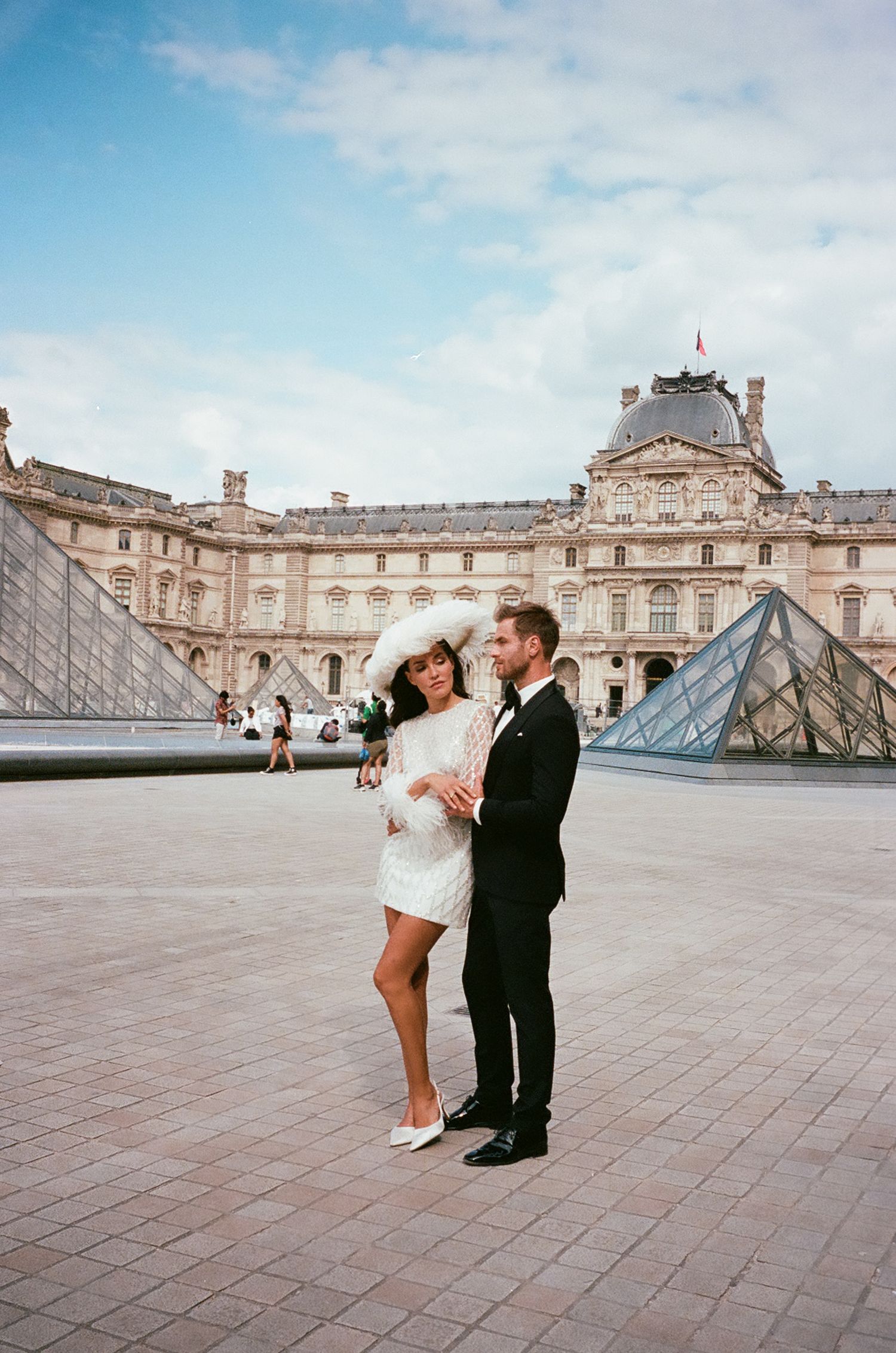 A couple poses together in front of the iconic glass pyramid and historic facade of the Louvre Museum in Paris.