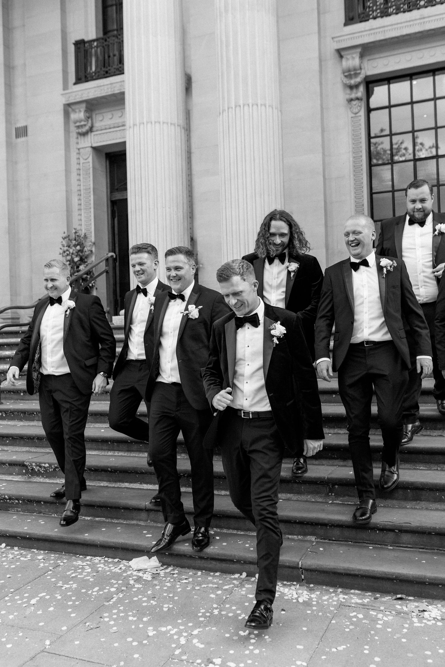 Groomsmen in black tuxedos lined up along classical building facade.