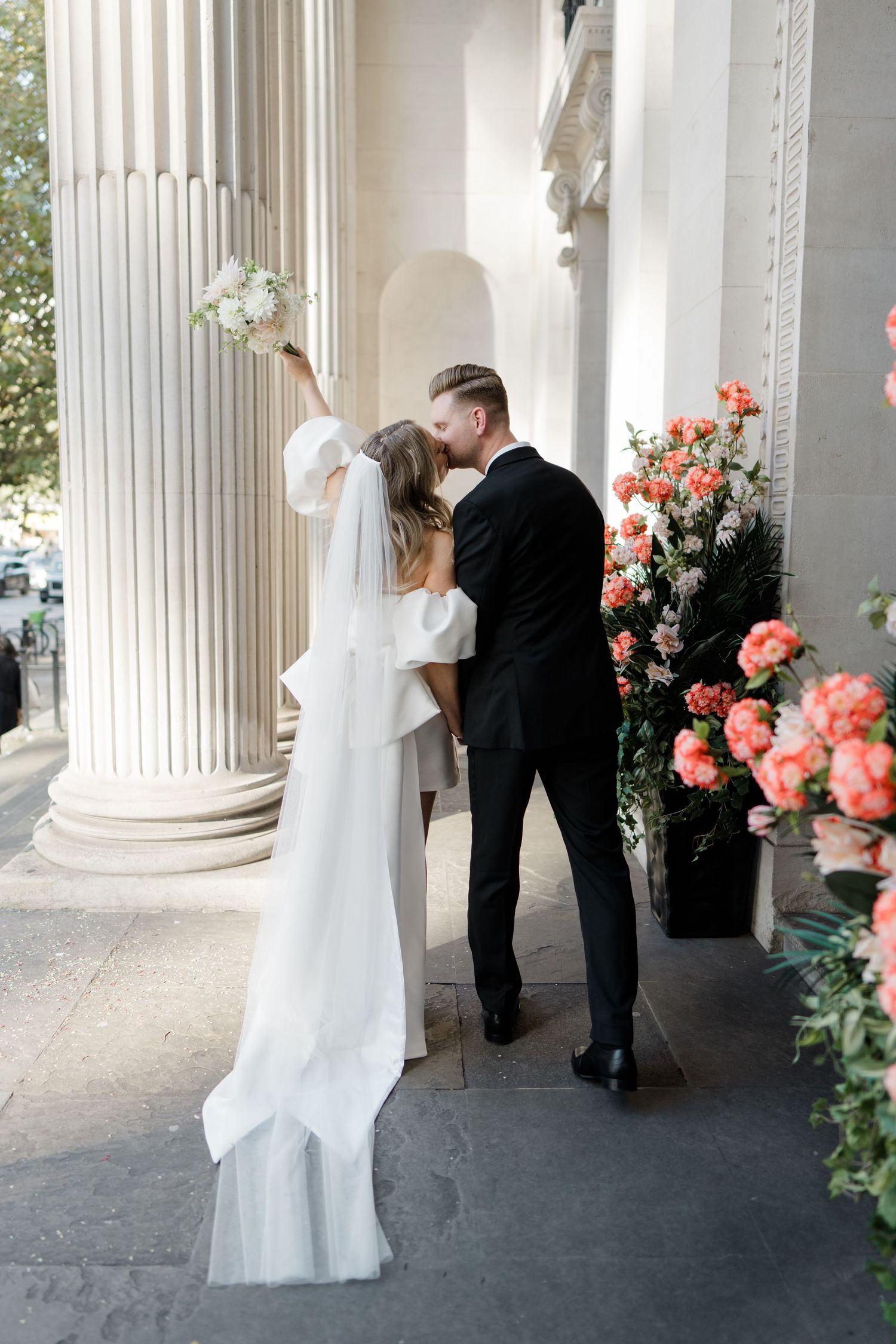 Wedding couple shares a moment under elegant white columns decorated with coral roses.