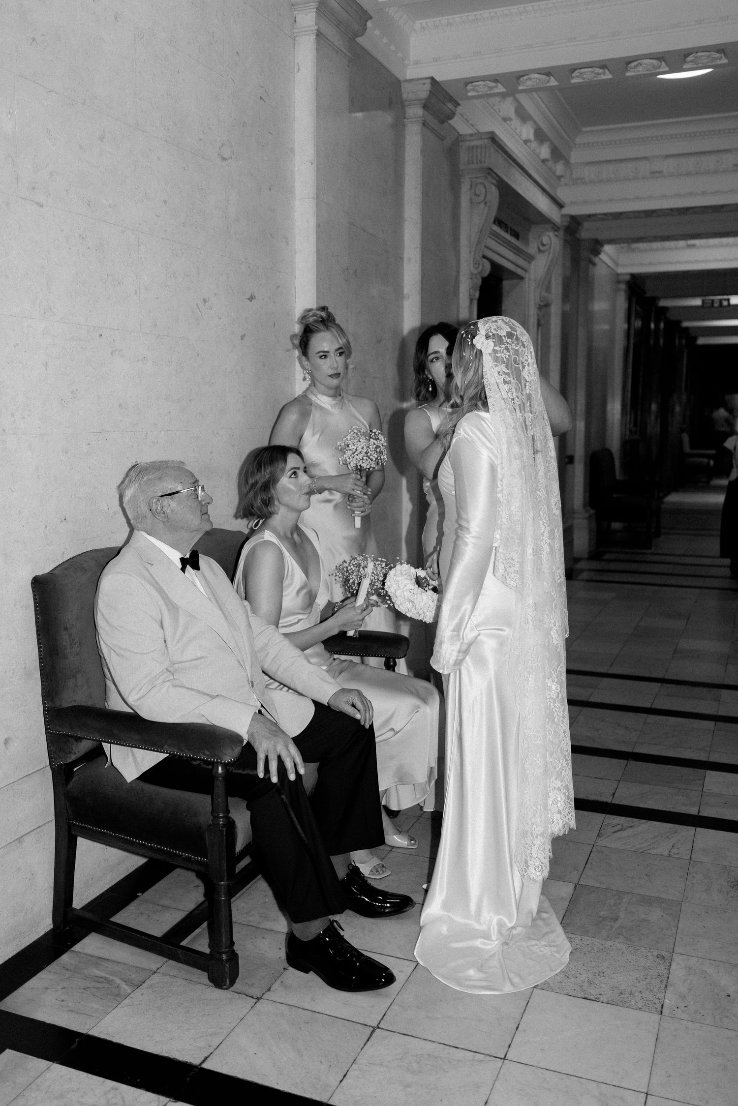 A wedding party takes a quiet moment in a grand hallway with classical architecture.