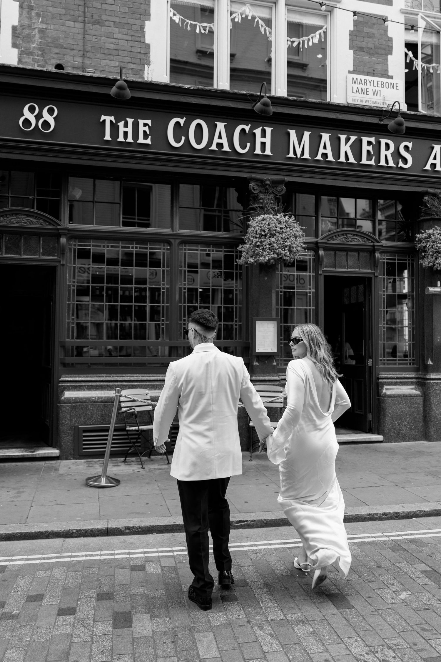 A couple walking away on a cobblestone street outside The Coach Makers pub in black and white.