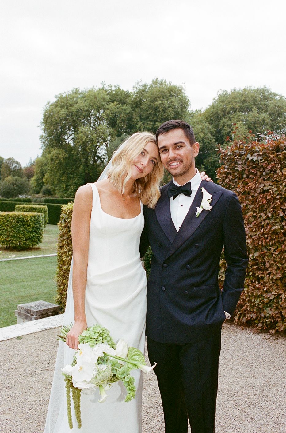 Bride in white gown holding bouquet poses with groom in navy tuxedo in lush garden setting.