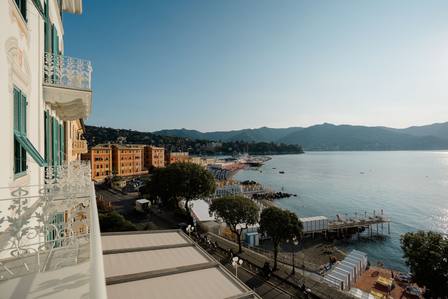 Scenic Italian Riviera coastal view from hotel balcony with turquoise bay, pier, and mountain backdrop.