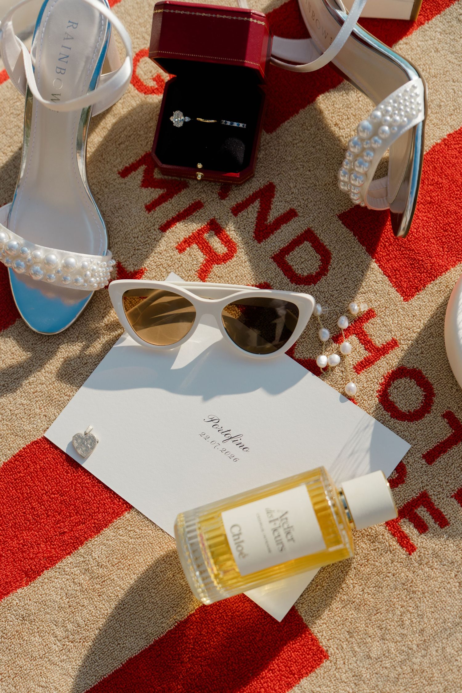 Bridal flat lay with pearl heels, cat-eye sunglasses, Cartier ring box, perfume bottle, and invitation on a striped towel.