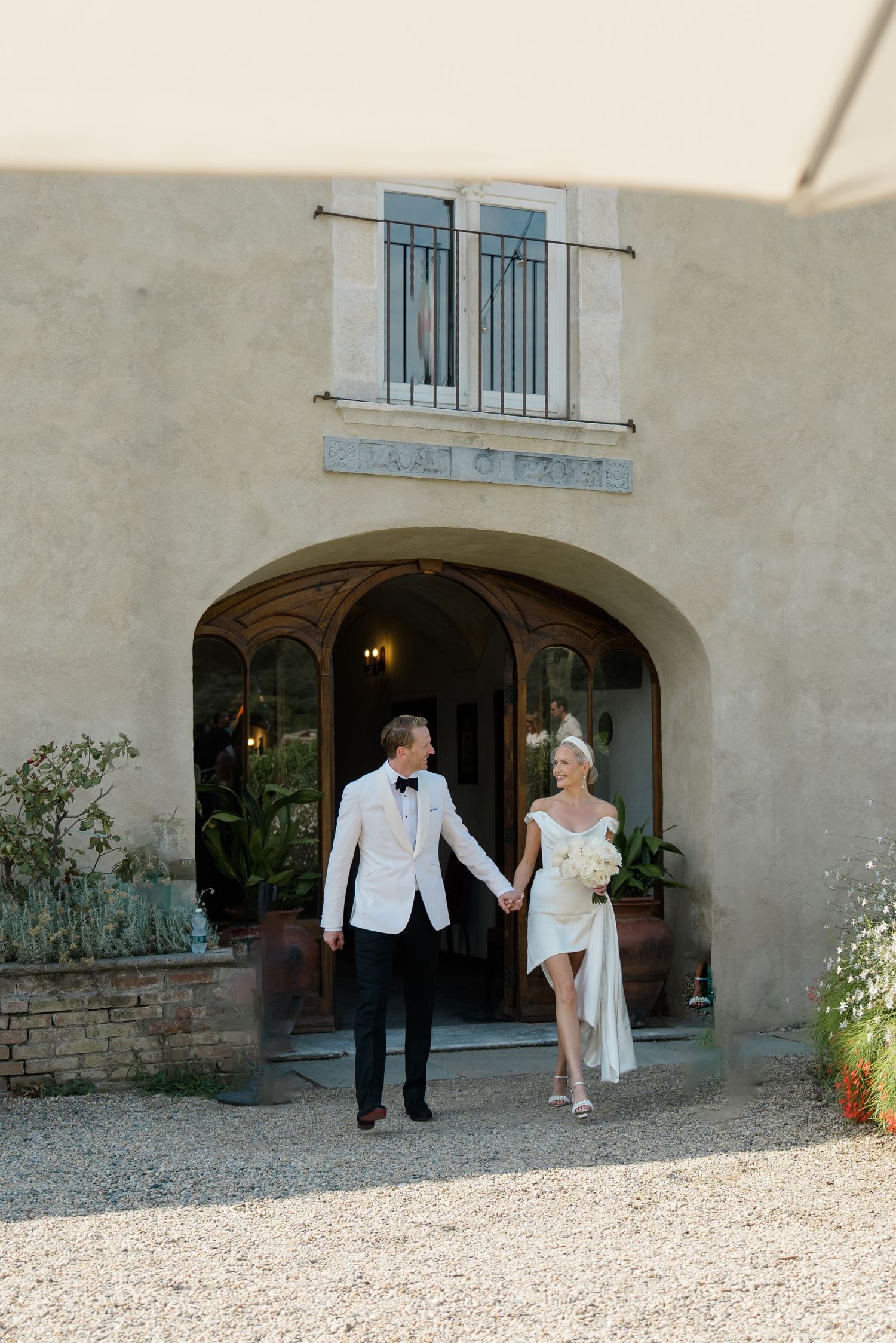 Newlywed couple holding hands walk out of a rustic Italian villa, bride in mini dress, groom in white tuxedo.