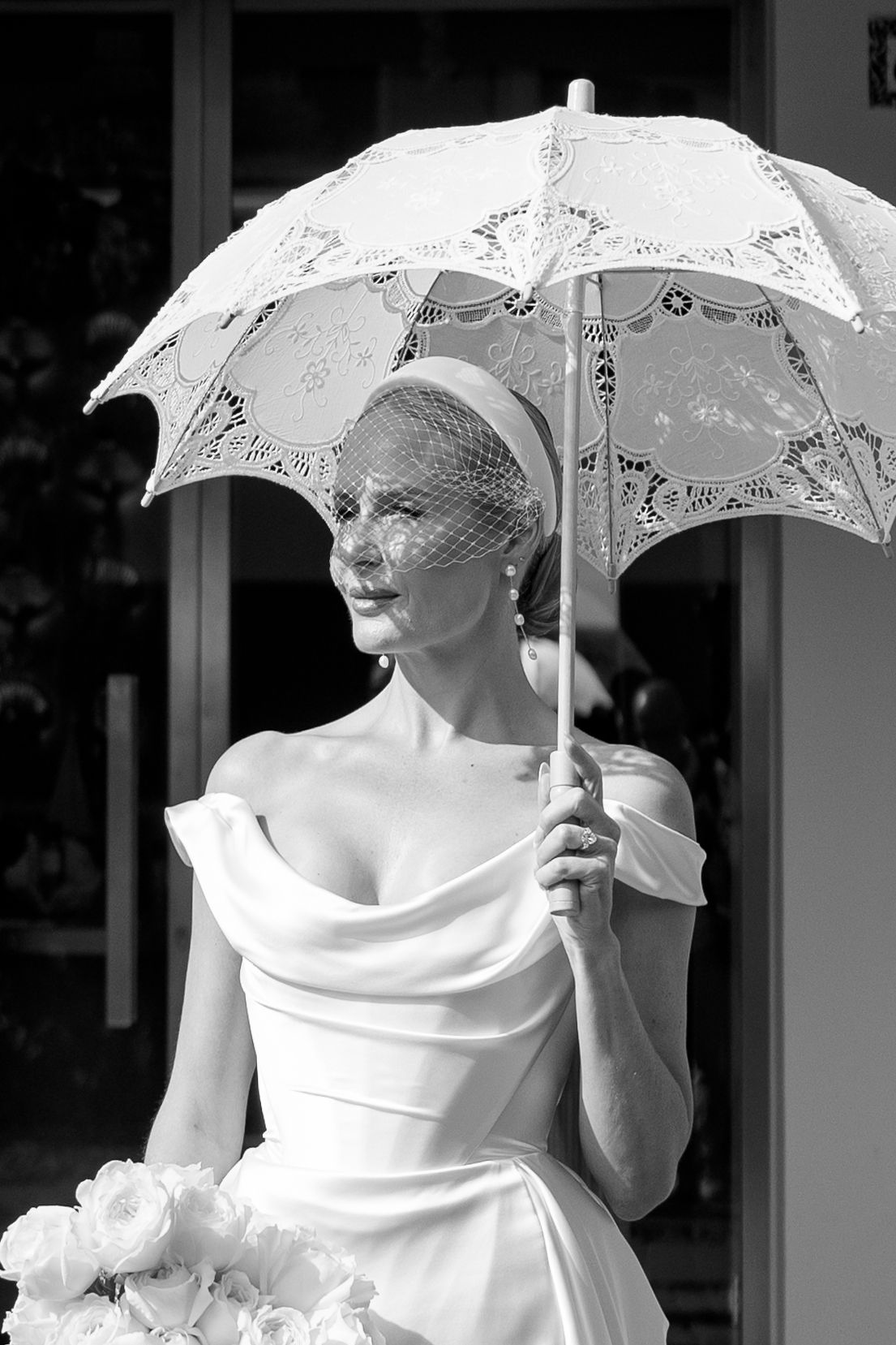 Black and white portrait of elegant bride in off-shoulder gown holding lace parasol with birdcage veil.