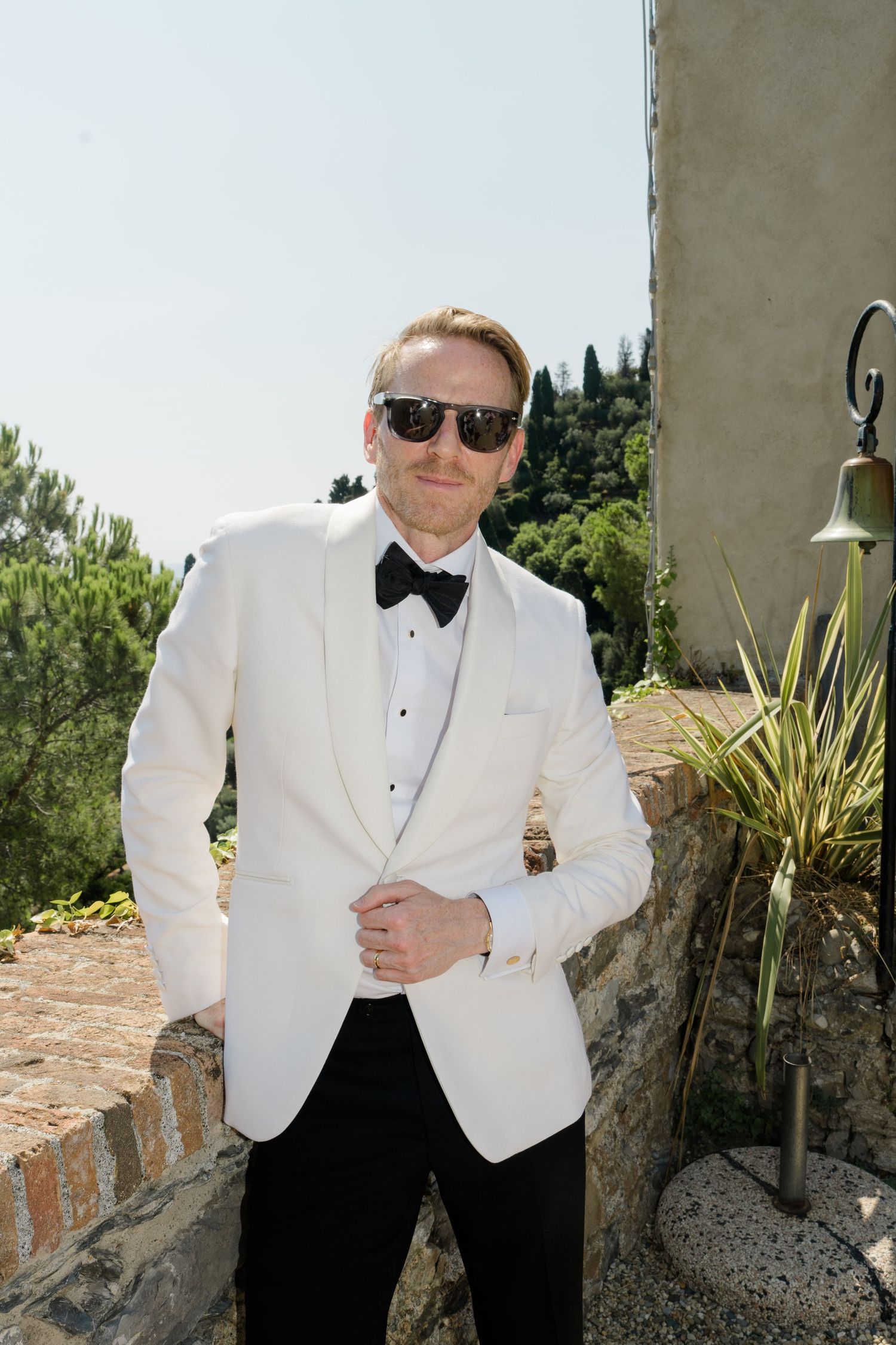 Dapper groom in white tuxedo jacket and black bow tie poses outdoors wearing sunglasses on sunny day.