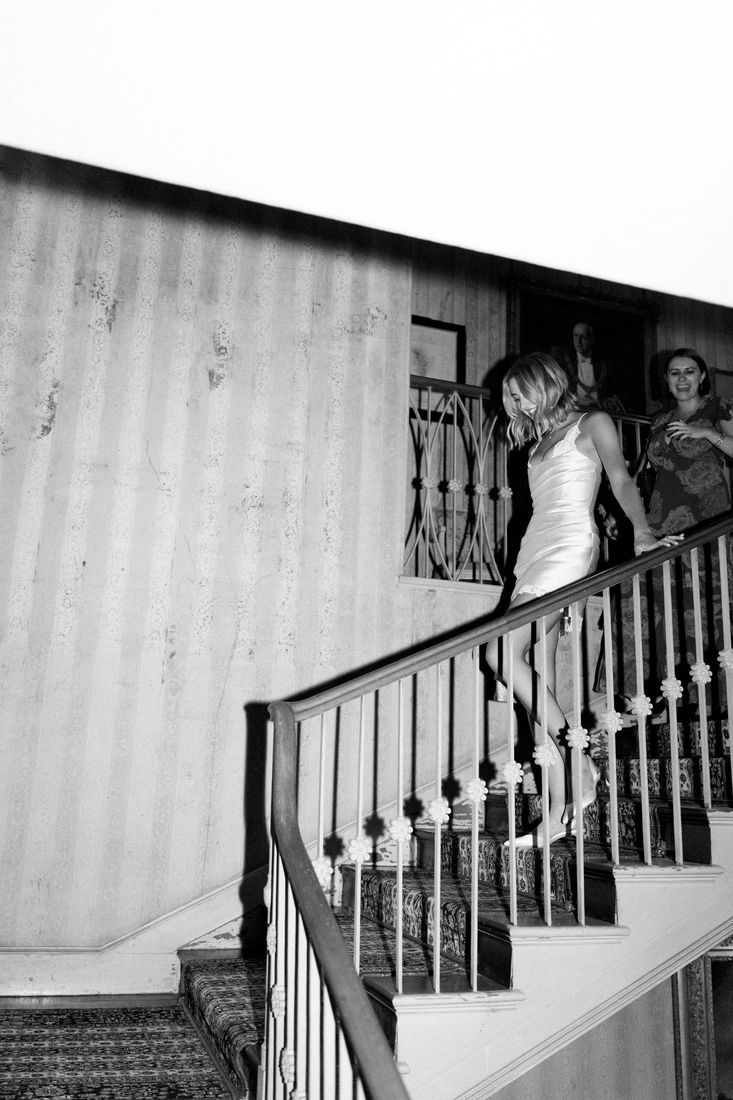 Bride in white dress descending decorated staircase in black and white wedding photo with guests watching.