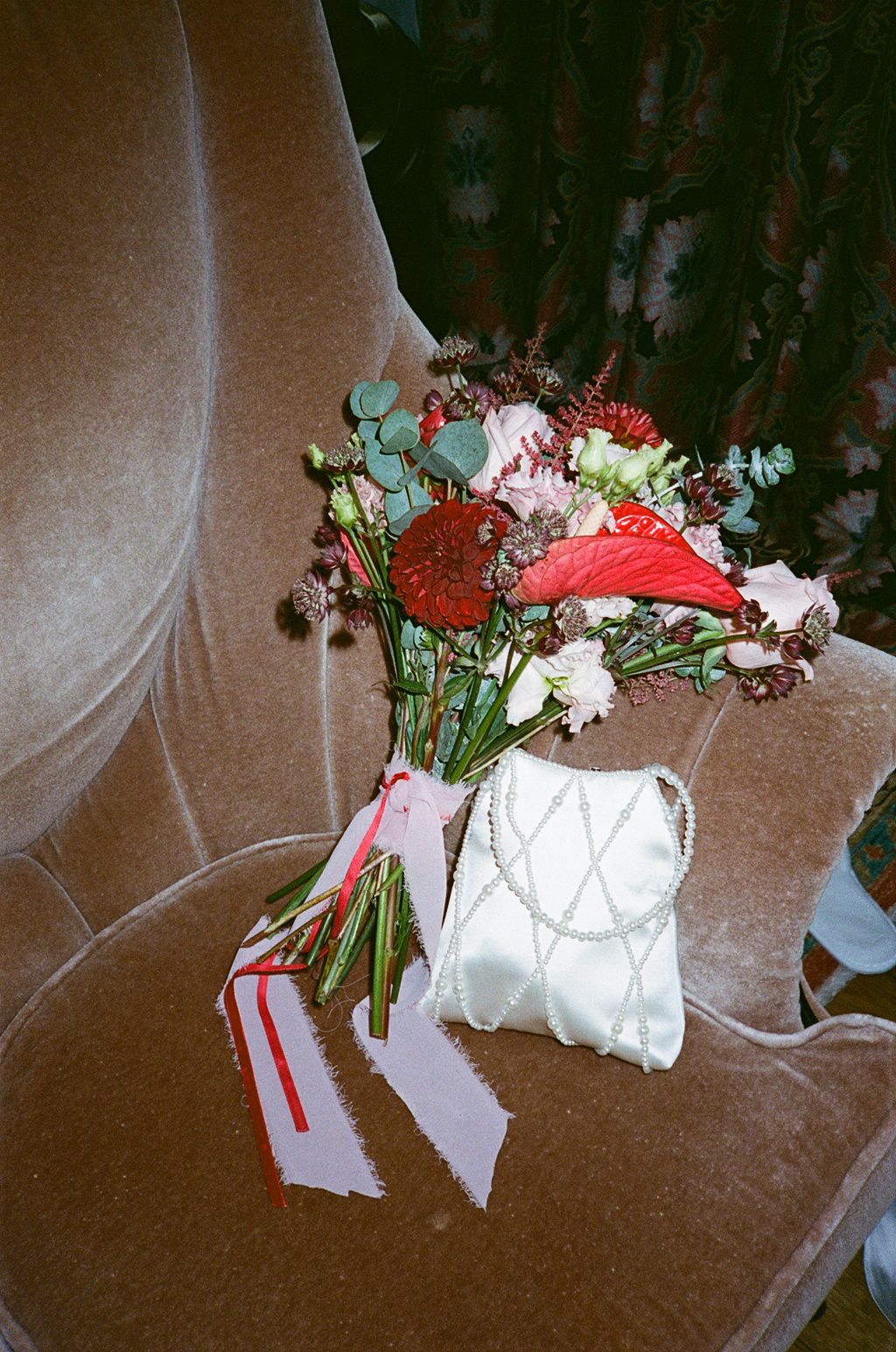 Red and pink floral bouquet with eucalyptus in a white beaded bag on a velvet chair.