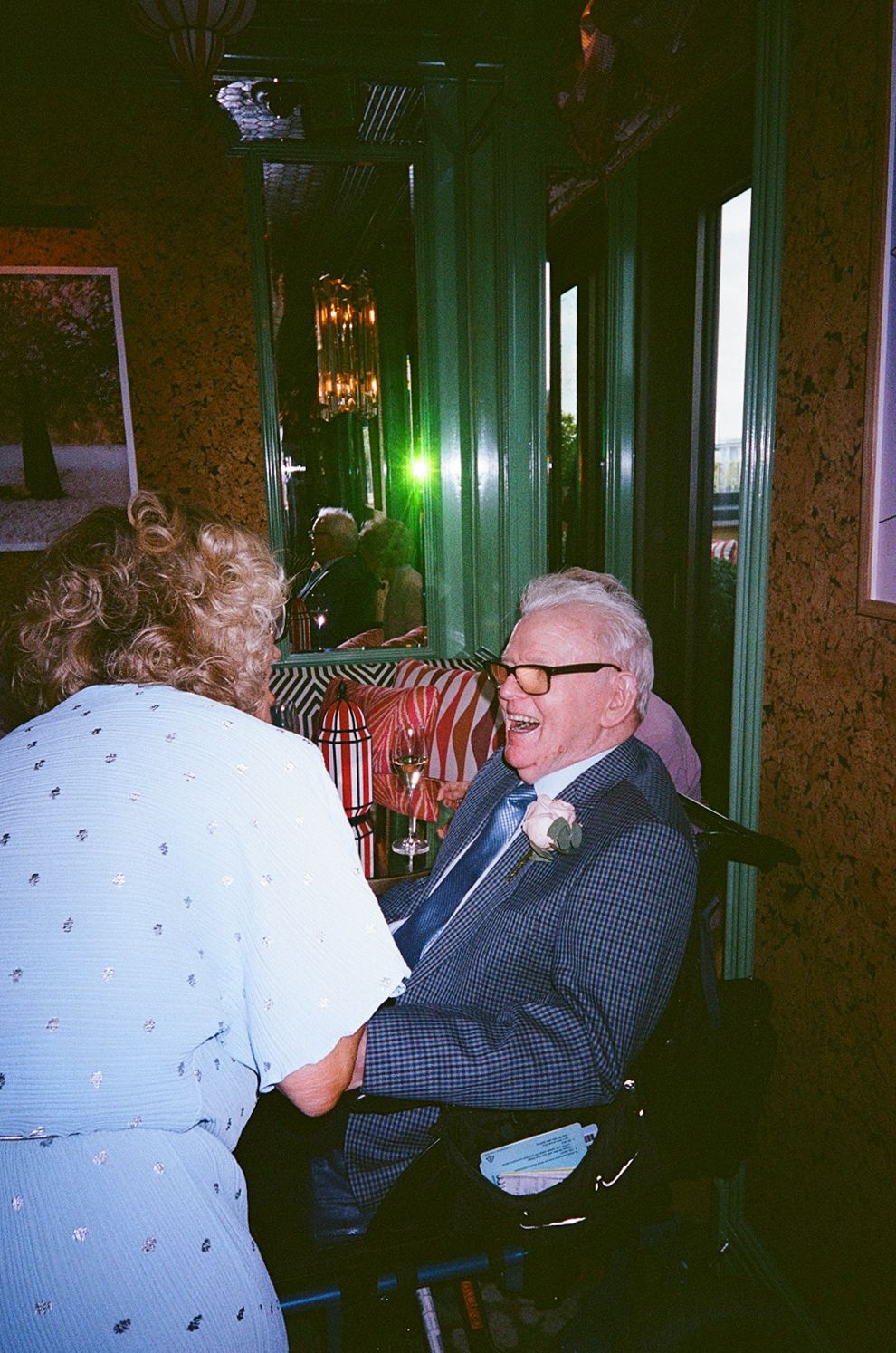 Elderly wedding guest in blue suit laughs joyfully with woman in ornate restaurant setting.