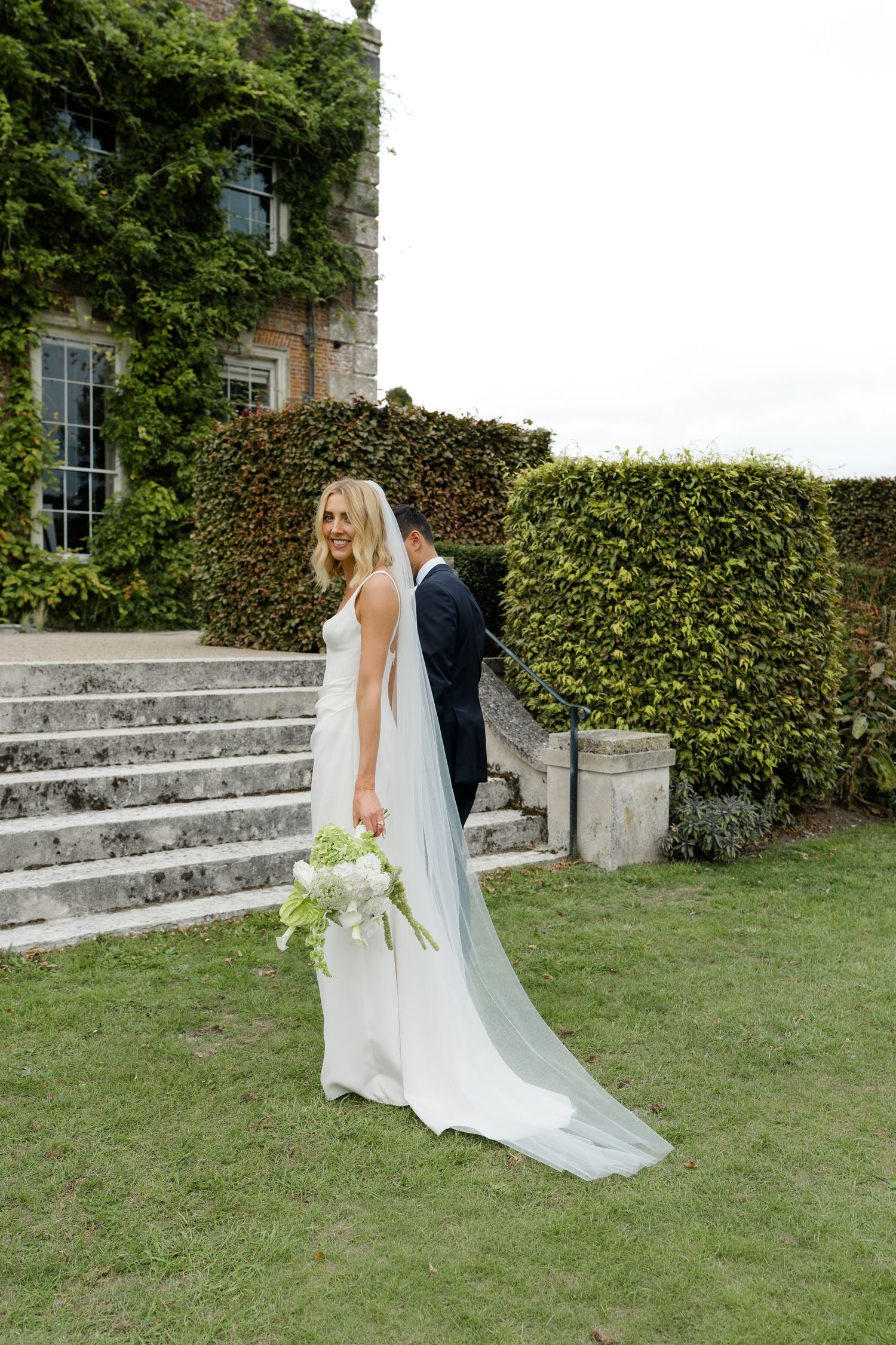 Bride in white gown with long veil outside ivy-covered estate holding bouquet.