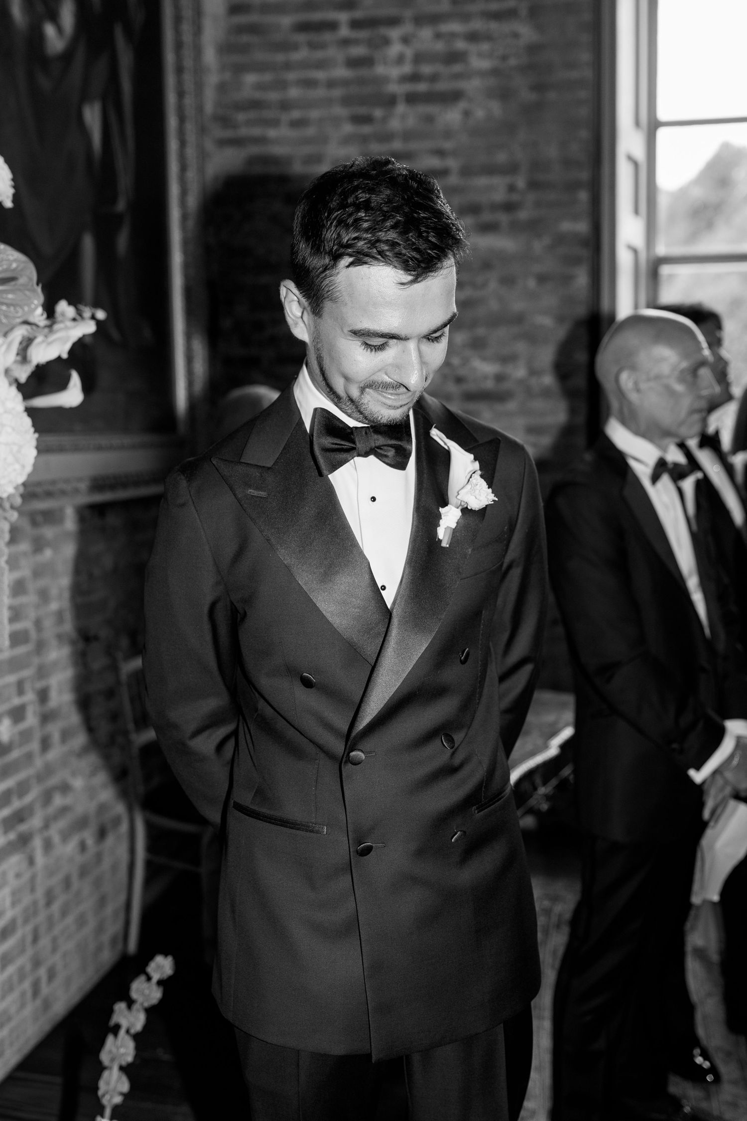 Groom in black tuxedo with bow tie and boutonniere looking down in black and white.