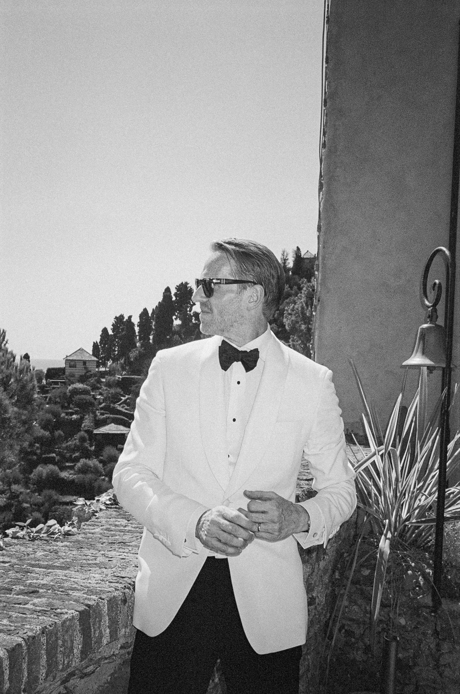 Groom in white tuxedo jacket and bow tie posing outdoors in black and white photo.