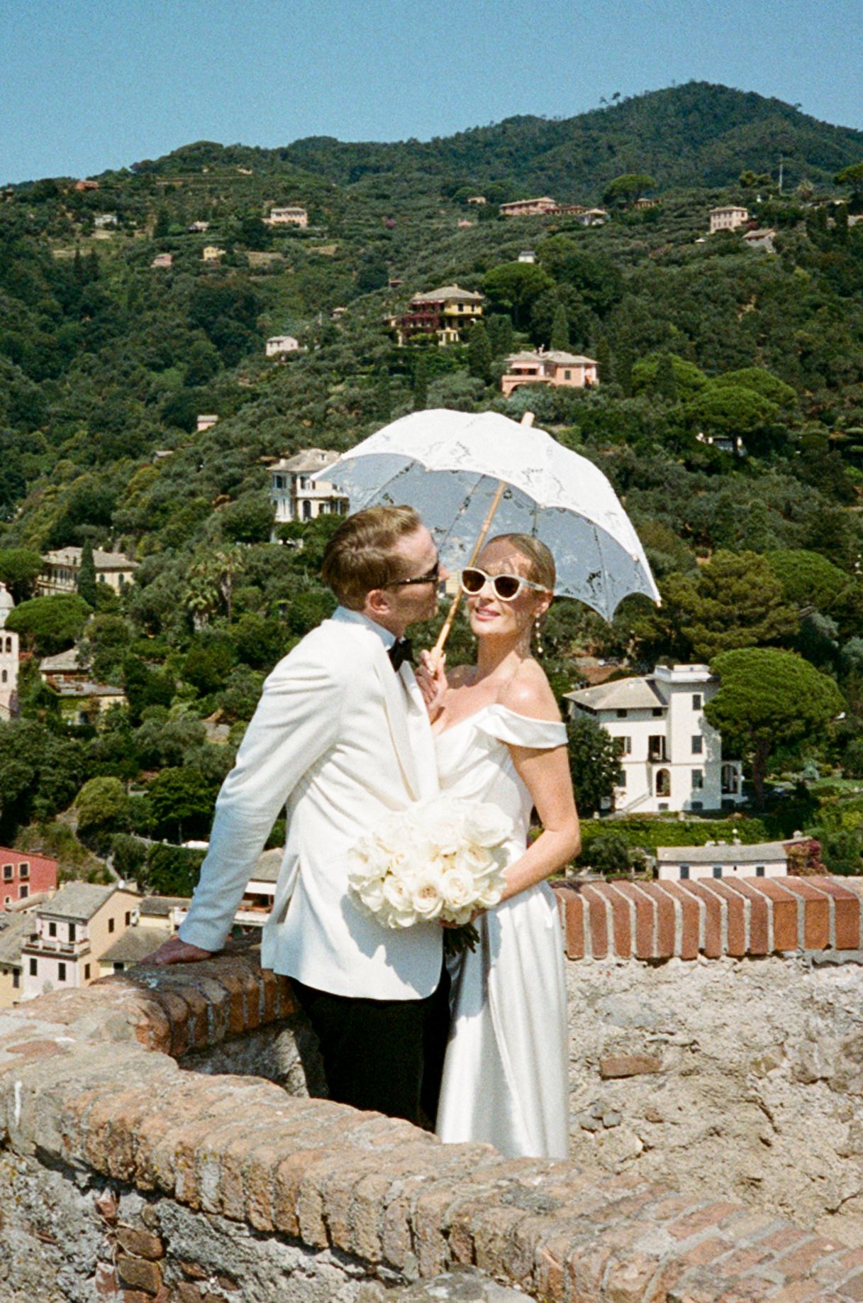 Bride and groom in white wedding attire pose with a lace parasol on a stone wall overlooking Portofino, Italy.