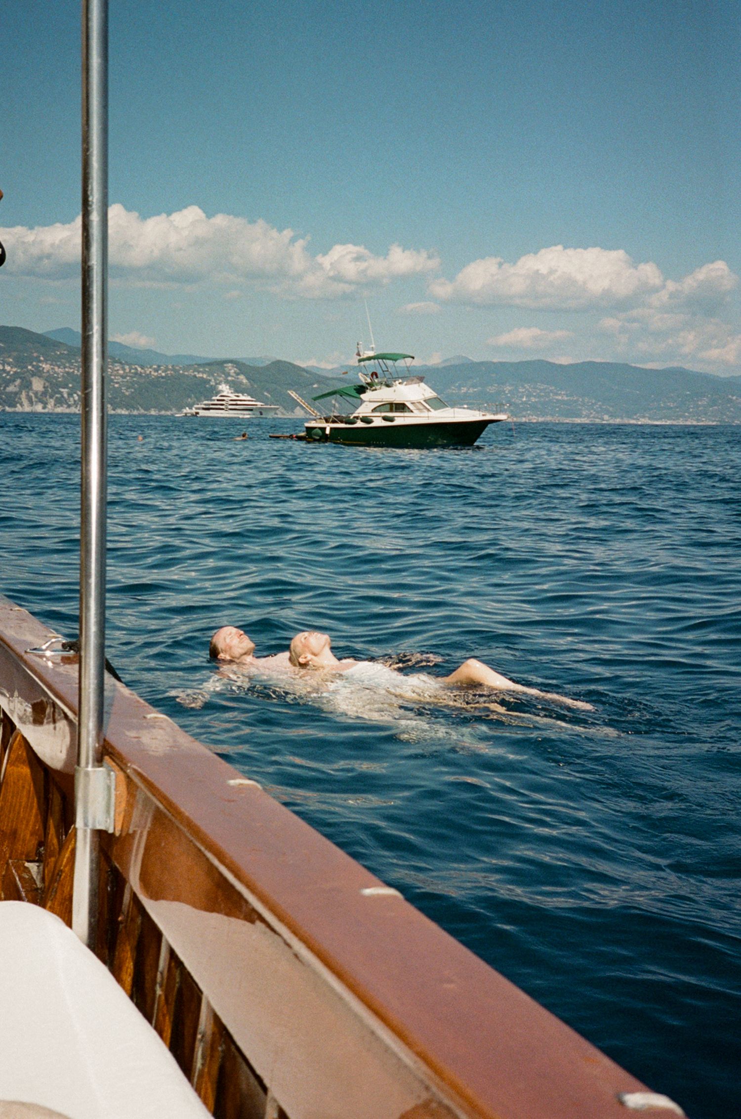 Newlyweds swimming in the blue Mediterranean Sea beside a classic wooden yacht on a sunny day.