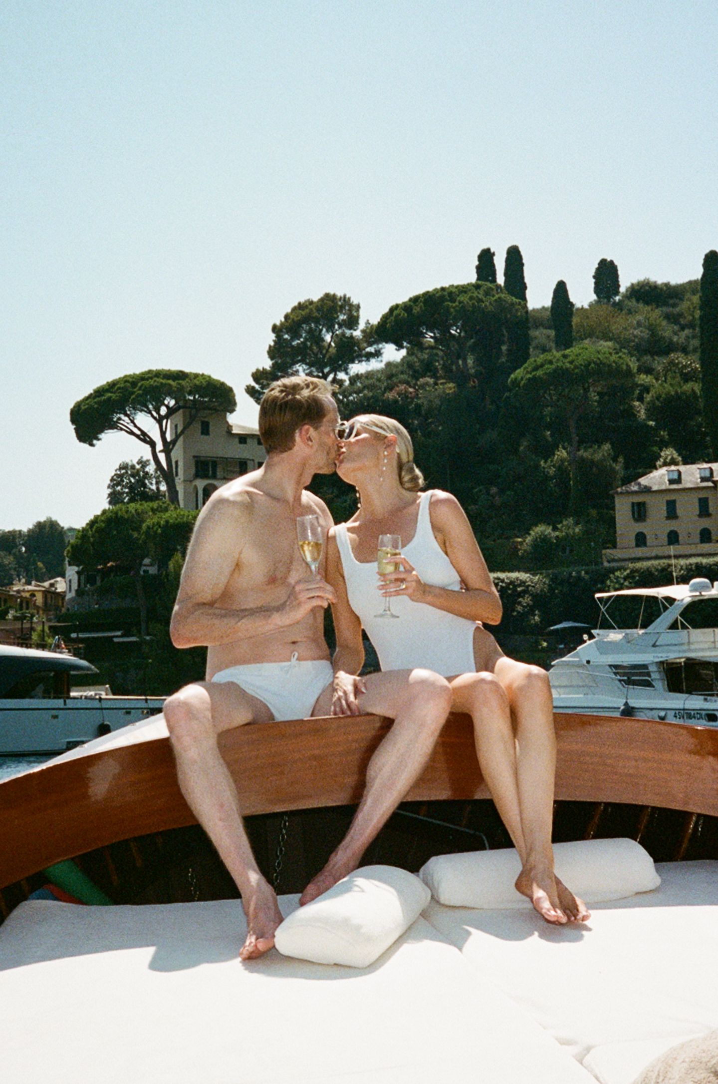 Newlyweds in white swimwear share a kiss while toasting champagne on a boat in an Italian harbor.