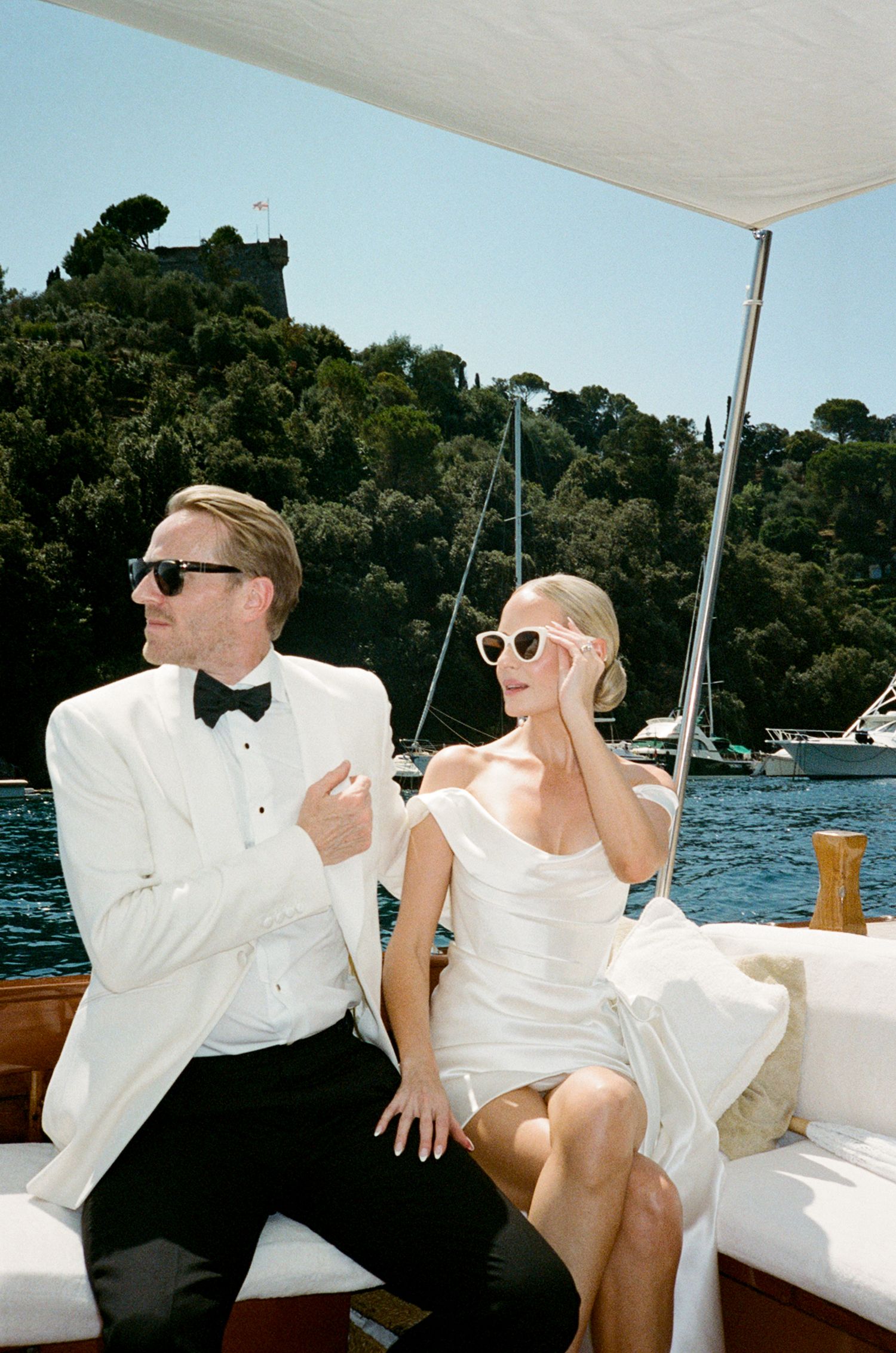 Stylish newlyweds in white wedding attire relax on a luxury wooden boat in a scenic Italian harbor.