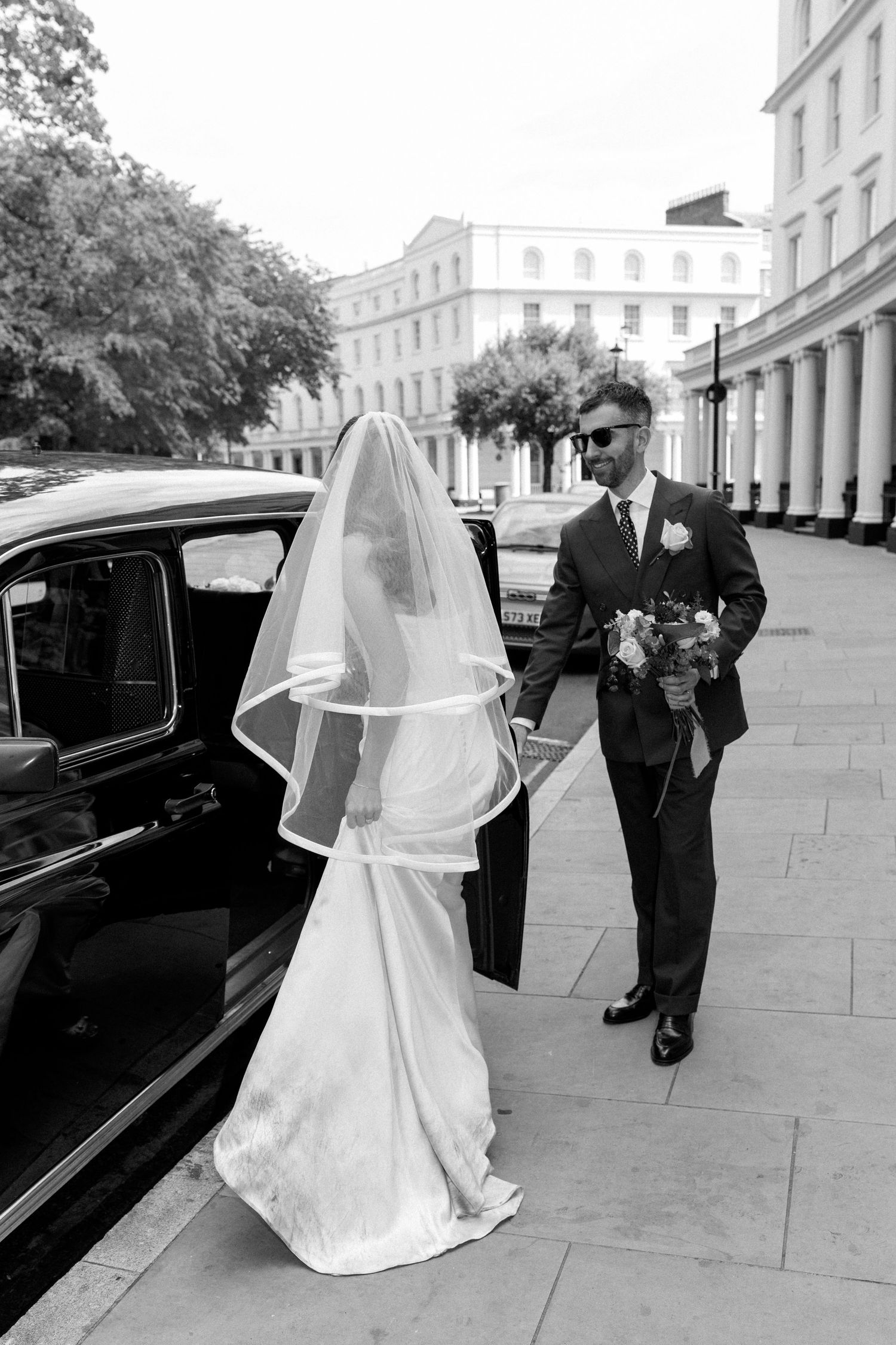 Bride in white gown and veil with groom holding bouquet beside vintage black car on elegant city street.