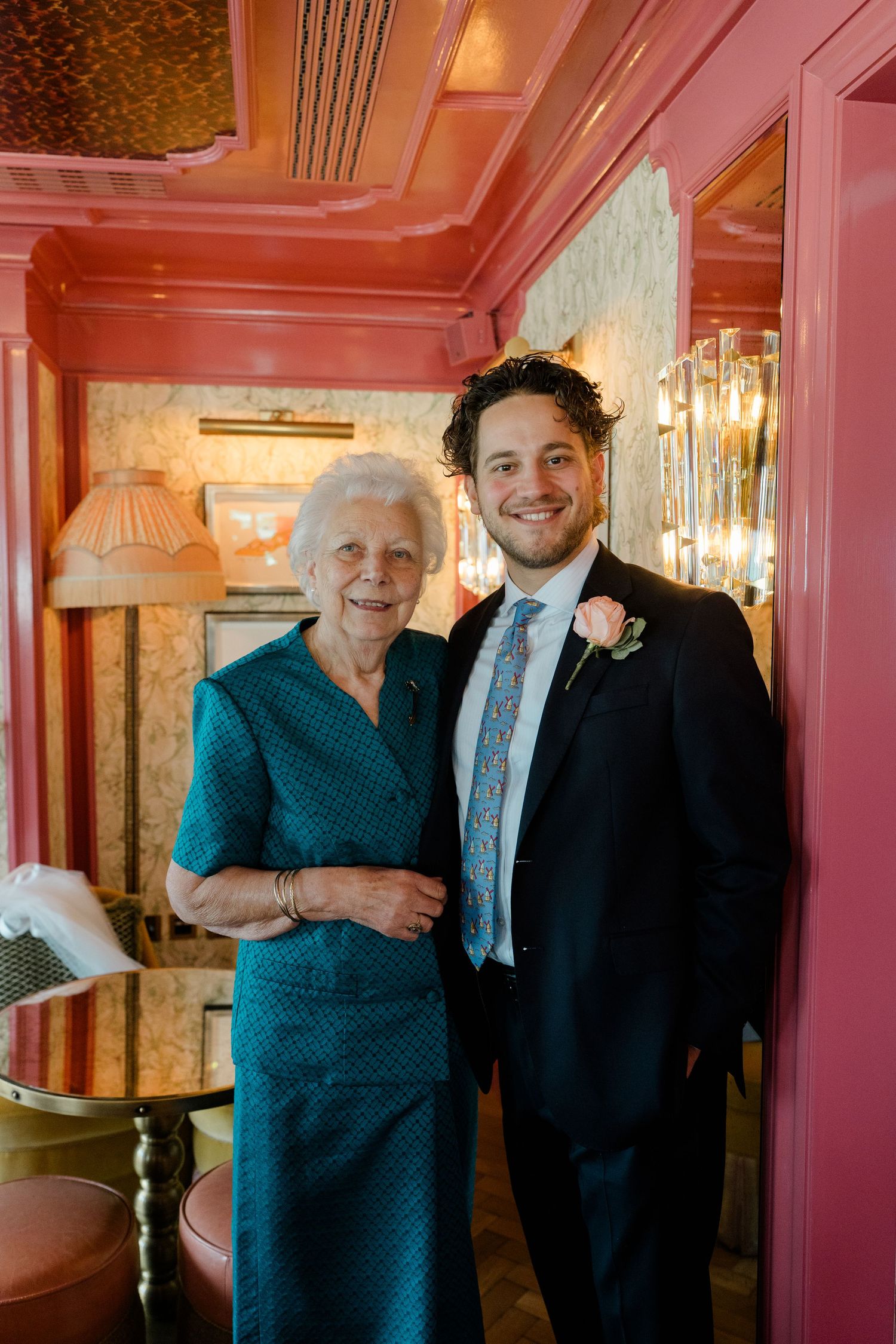 Young groom in dark suit with boutonniere posing with elderly woman in teal dress at wedding venue.