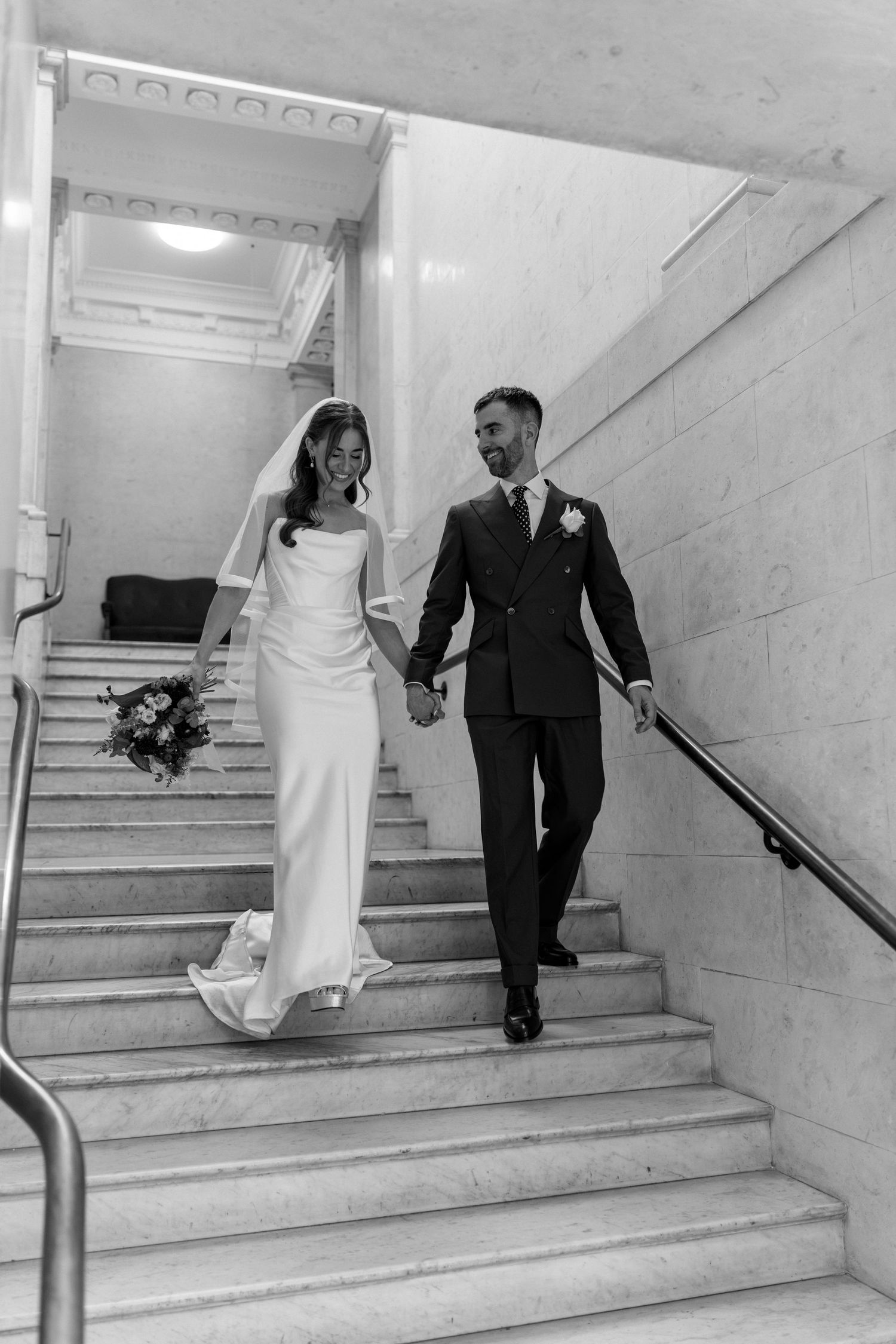 Newlyweds holding hands and smiling as they walk down marble stairs after their ceremony.