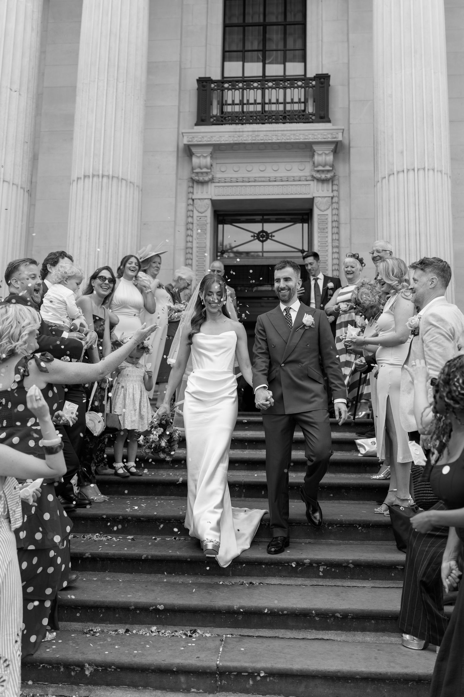 Newlyweds walk hand in hand through confetti thrown by guests on grand building steps.