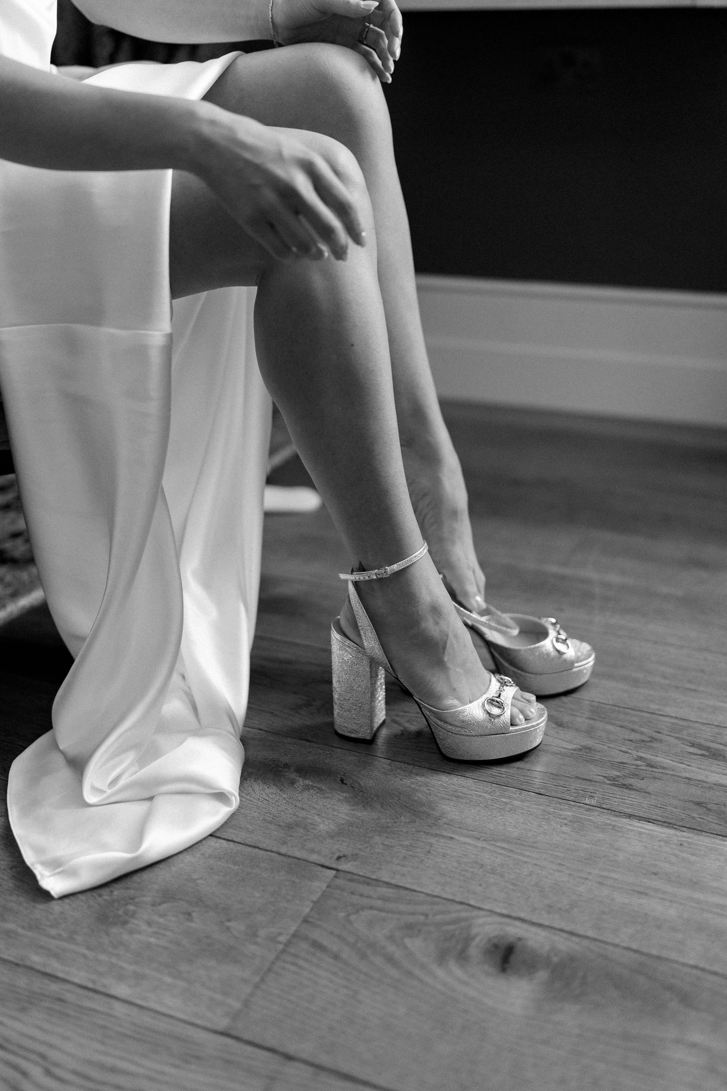 Black and white photo of a bride fastening her strappy glitter platform heels while seated in her white wedding gown.