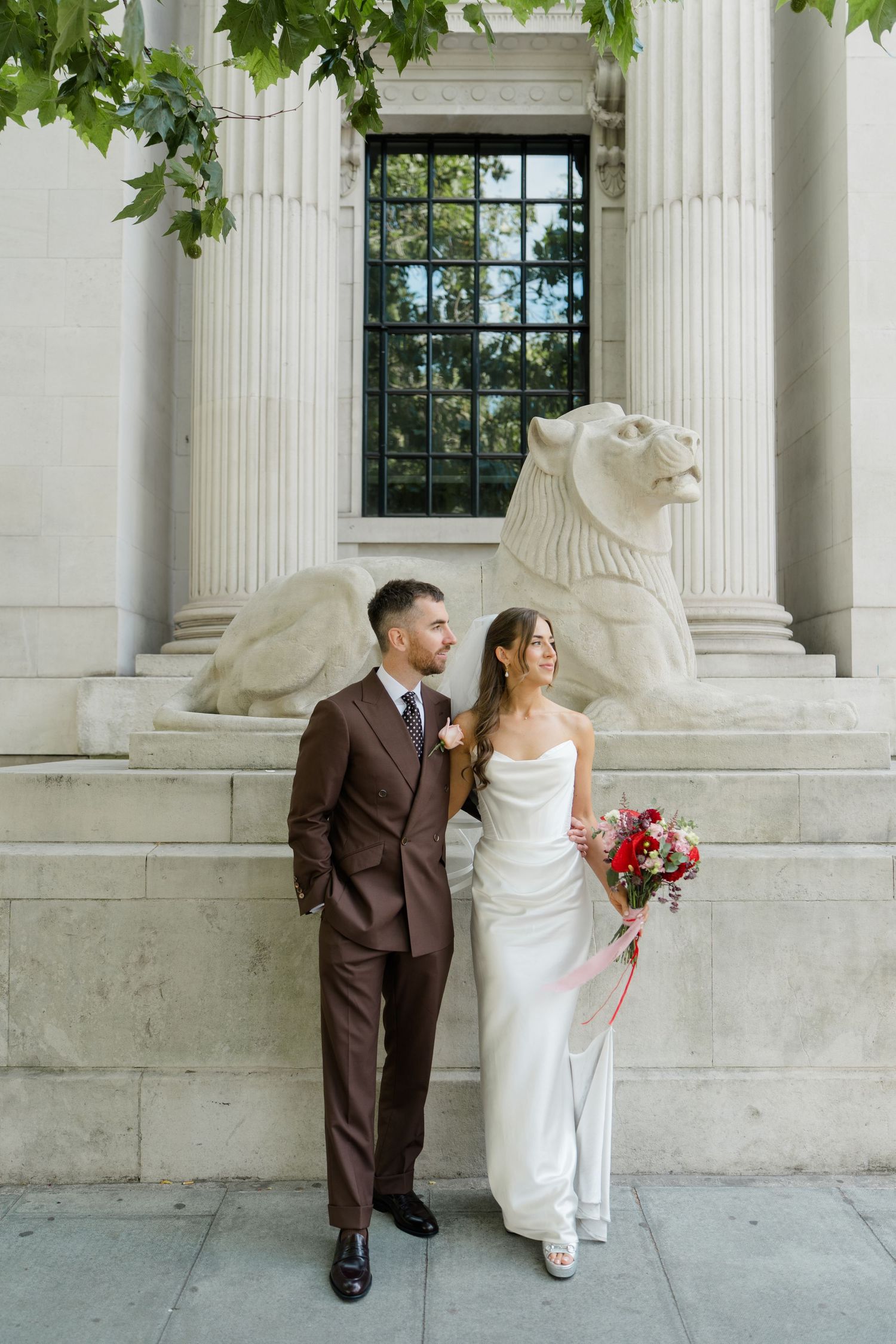 Bride and groom standing before stone lion sculpture and classical columns, bride holding vibrant red and pink bouquet.