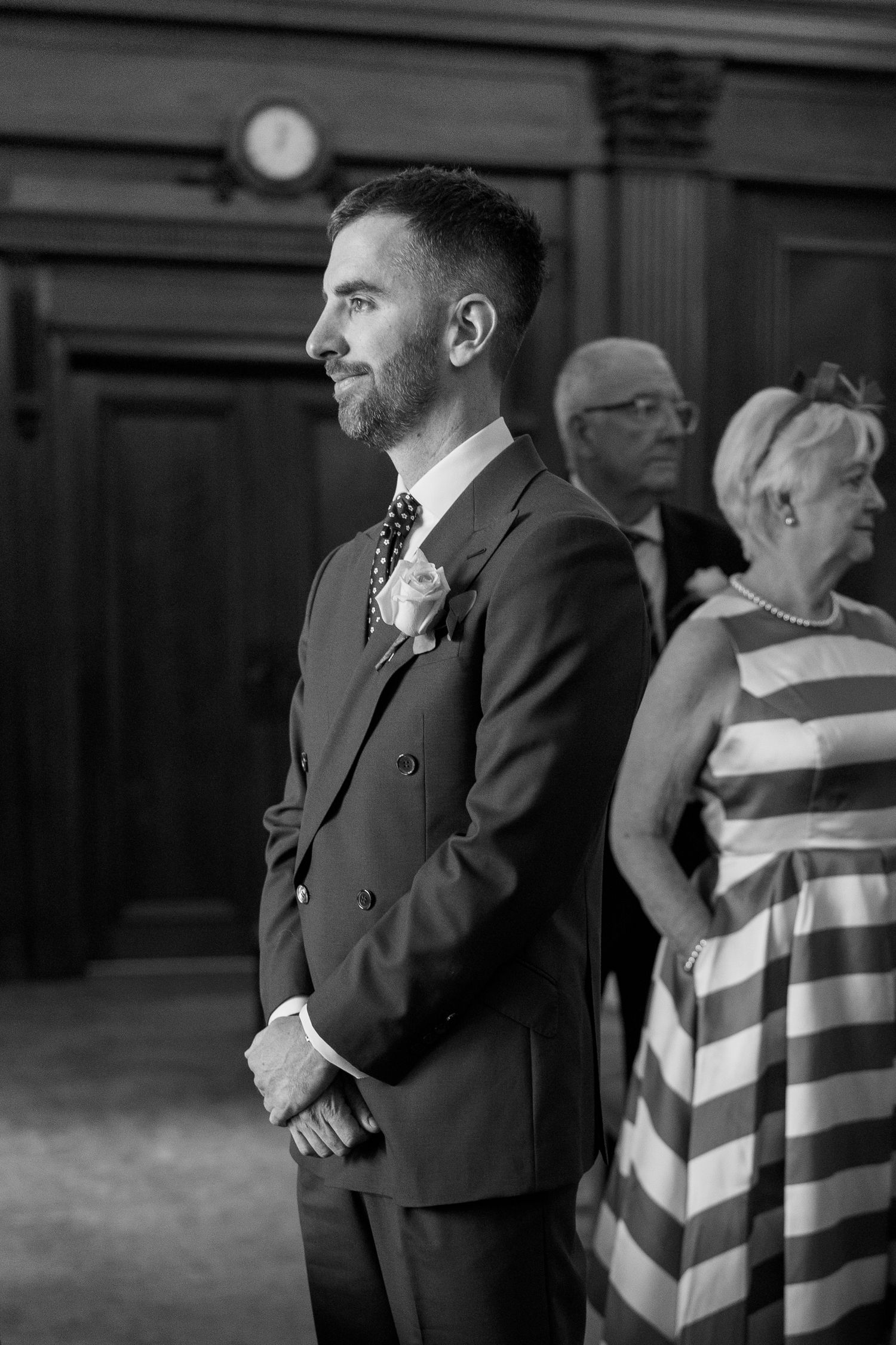 Groom in dark suit with boutonniere waiting at the altar with guests behind him.