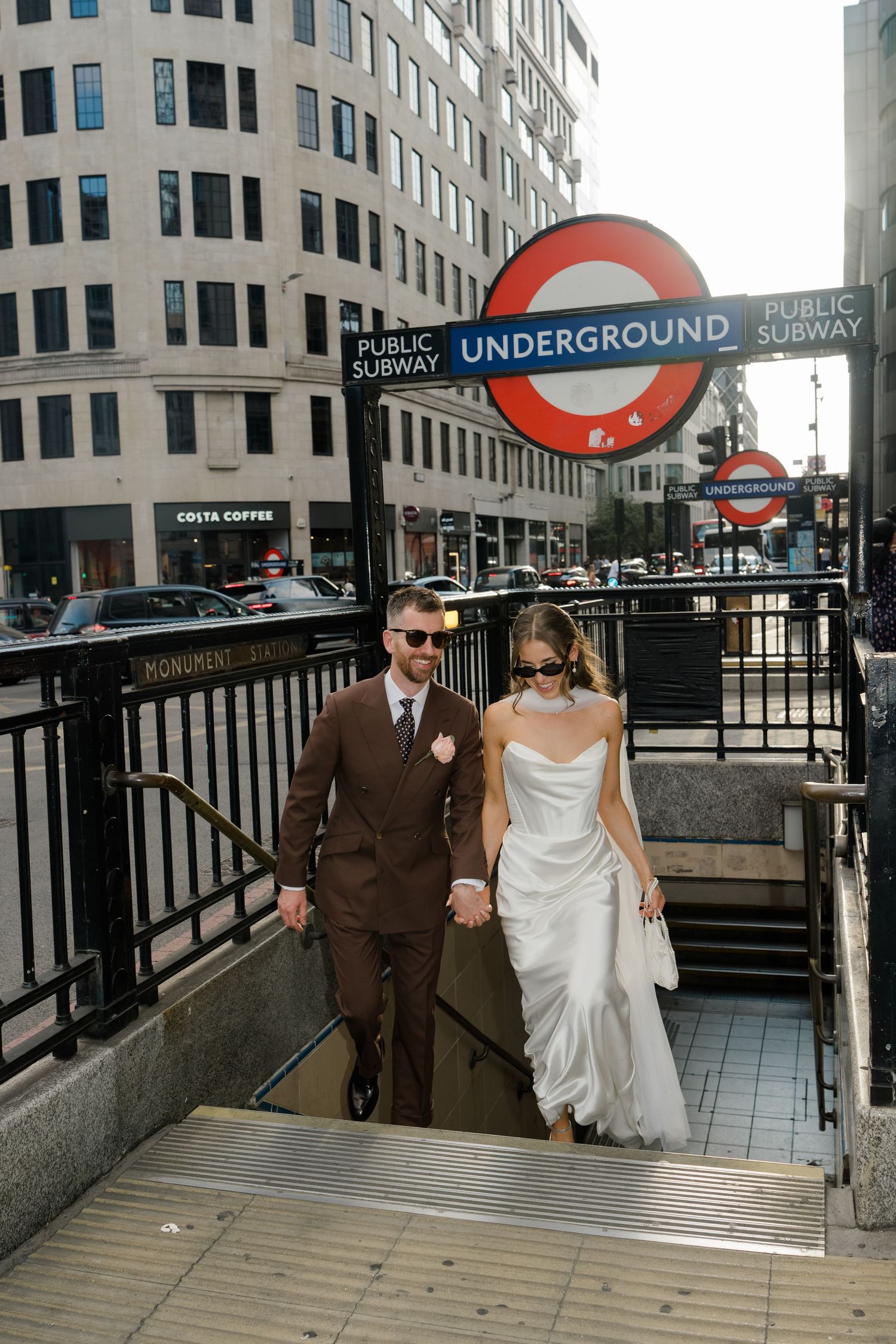 Bride and groom in wedding attire holding hands emerging from London Underground Monument station entrance.