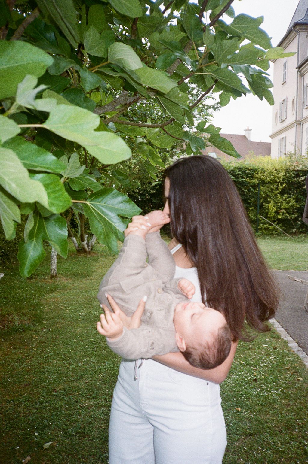 Young woman with long dark hair holding a baby outdoors near a fig tree in a lush green garden.