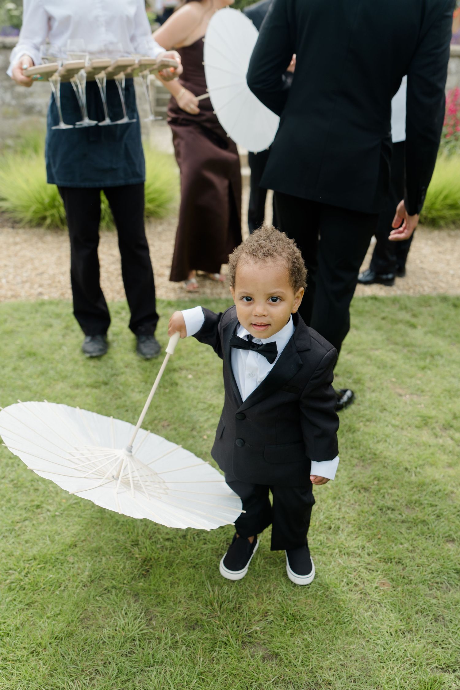 Adorable toddler in black tuxedo holding white parasol at outdoor wedding reception on green grass.