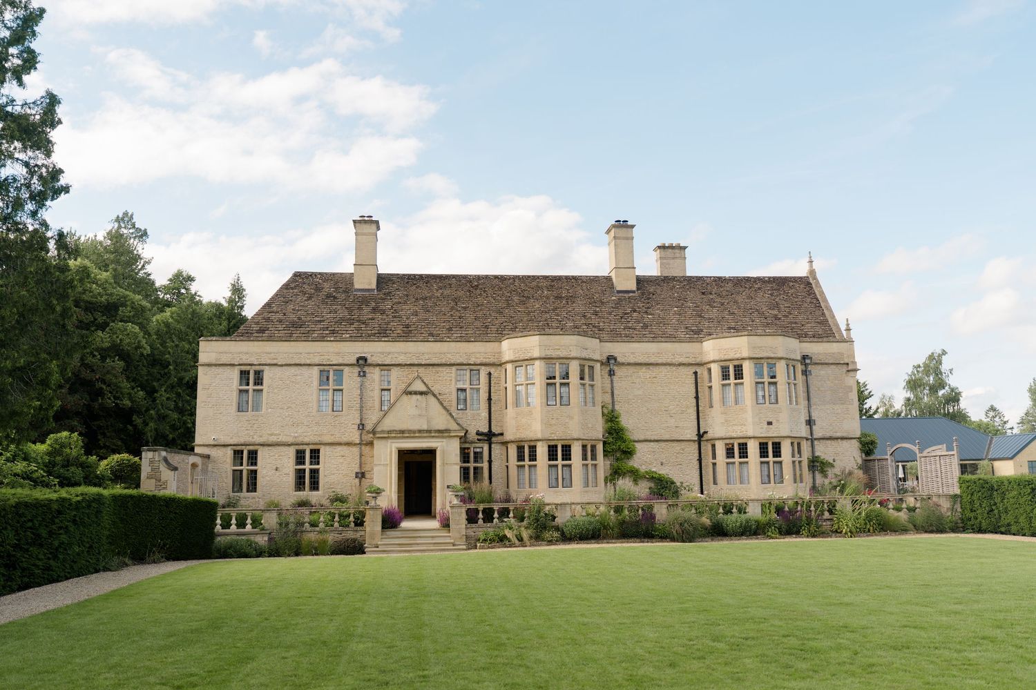 Historic Cotswold stone manor house with manicured lawn, tall chimneys, and lush green hedges under a blue sky.