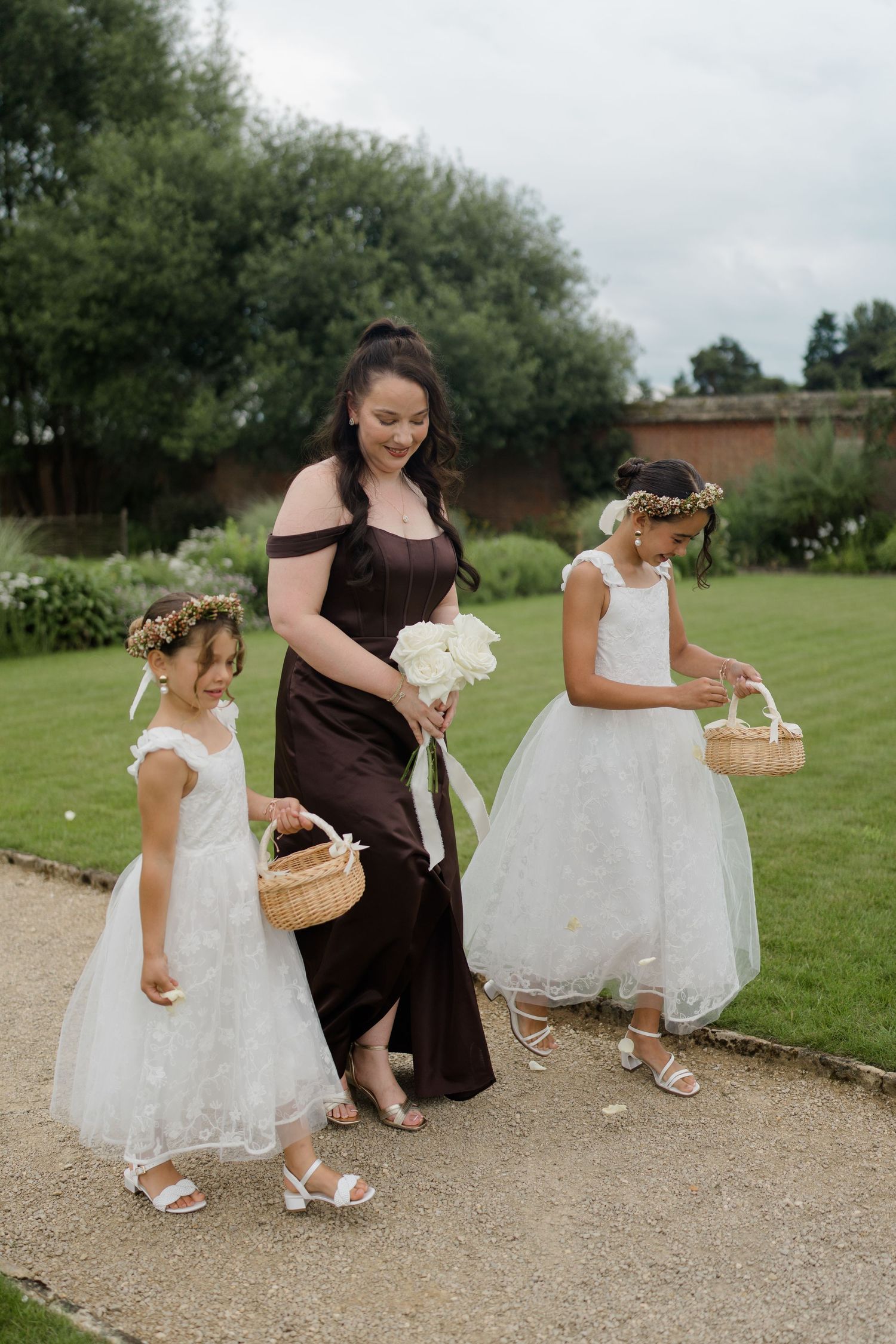 A bridesmaid in a dark brown gown walks with two young flower girls in white dresses carrying baskets.