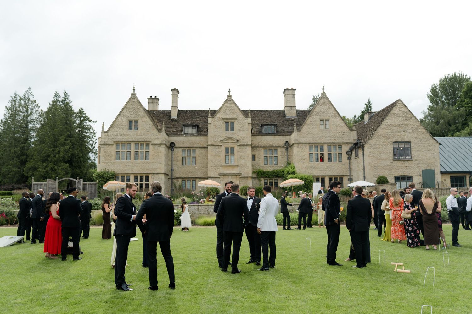 Wedding guests mingling on manicured lawn in front of grand English manor house during outdoor cocktail reception.