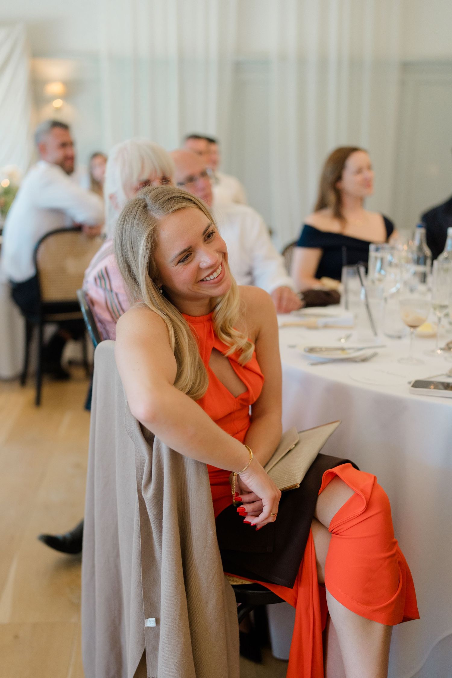 Smiling blonde woman in orange dress enjoys wedding reception at elegantly set dining table.