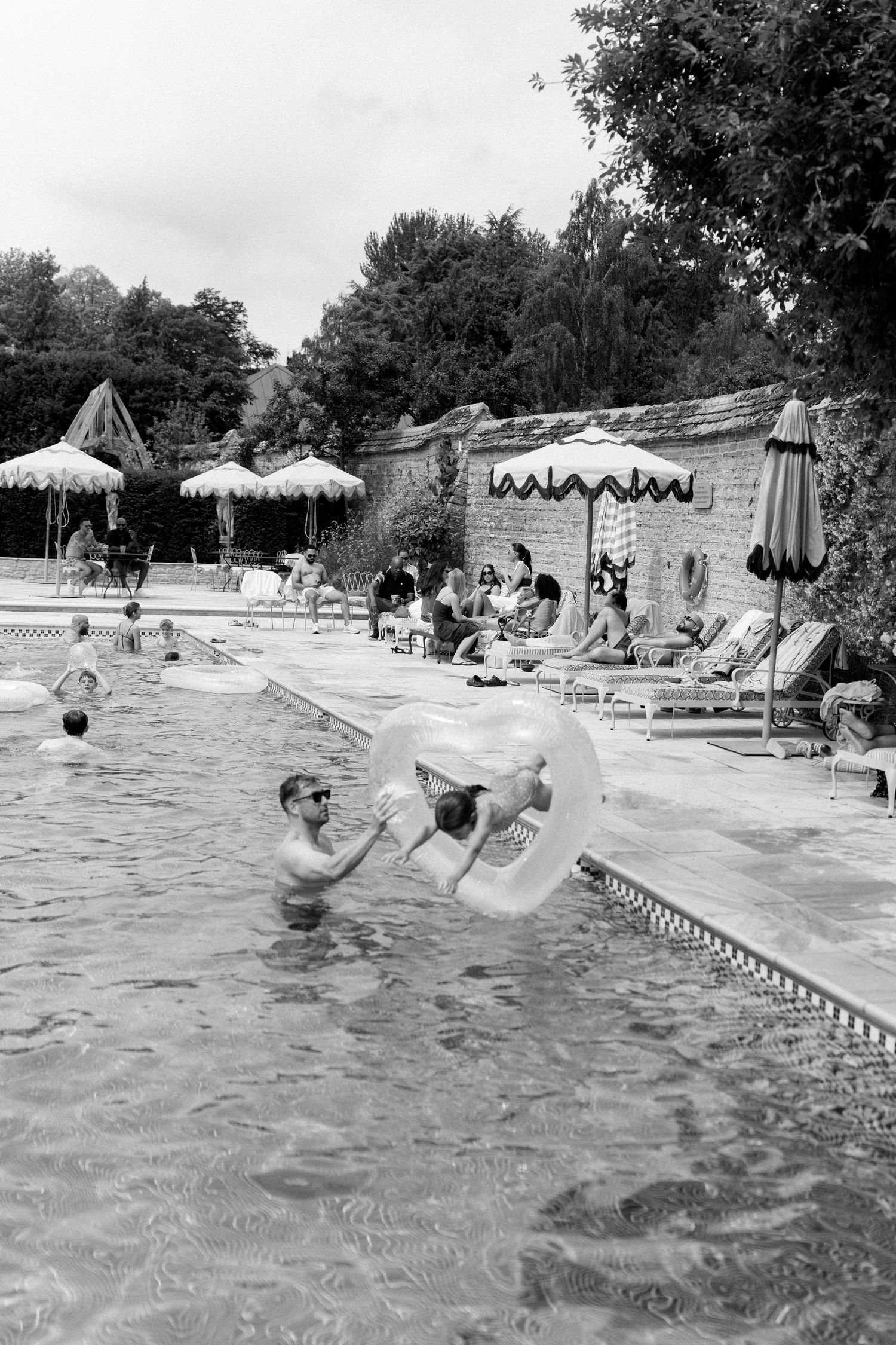 Black and white photo of man holding large clear inflatable ring in outdoor pool at wedding.