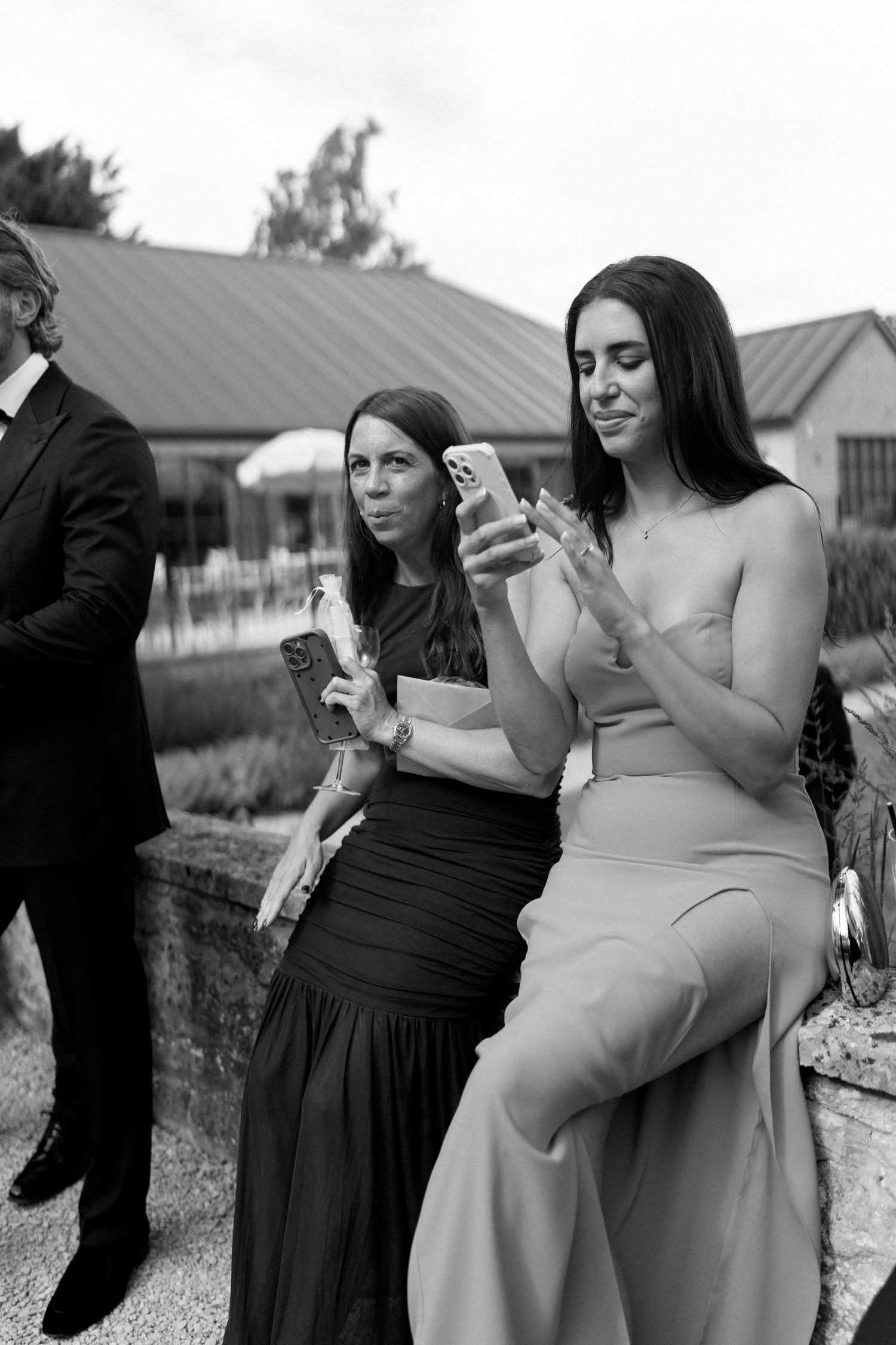 Black and white photo of two women in formal dresses looking at phones at outdoor wedding.