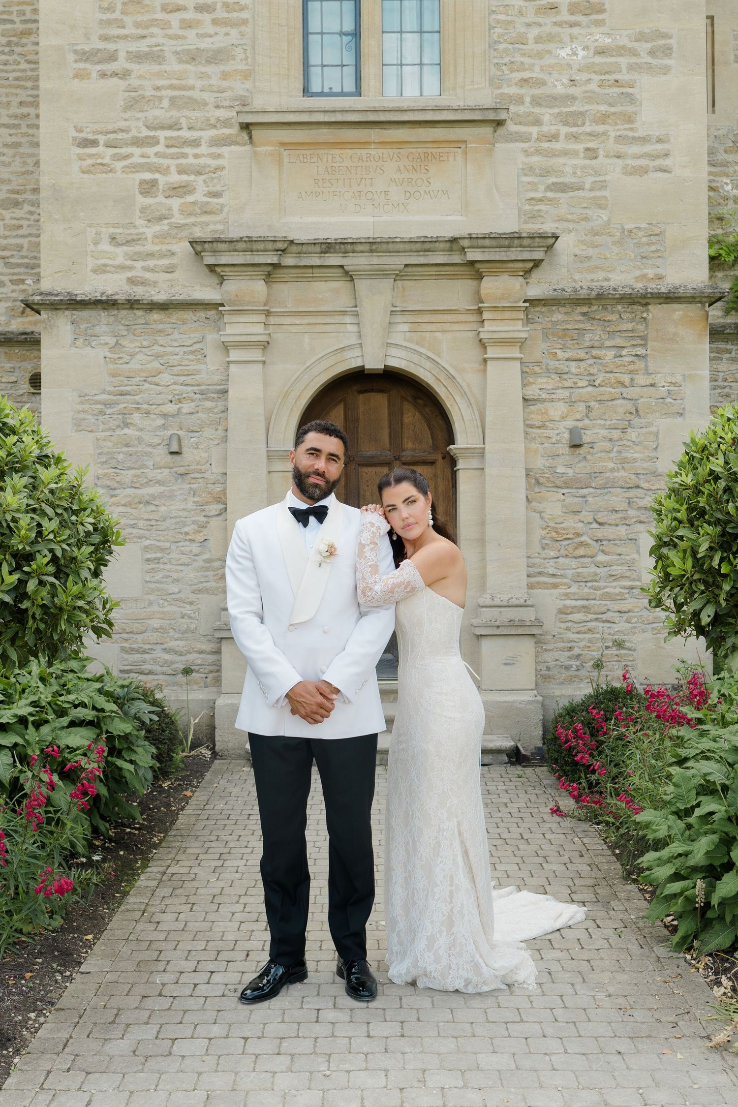 Bride and groom pose before ornate stone archway doorway surrounded by lush greenery at wedding venue.