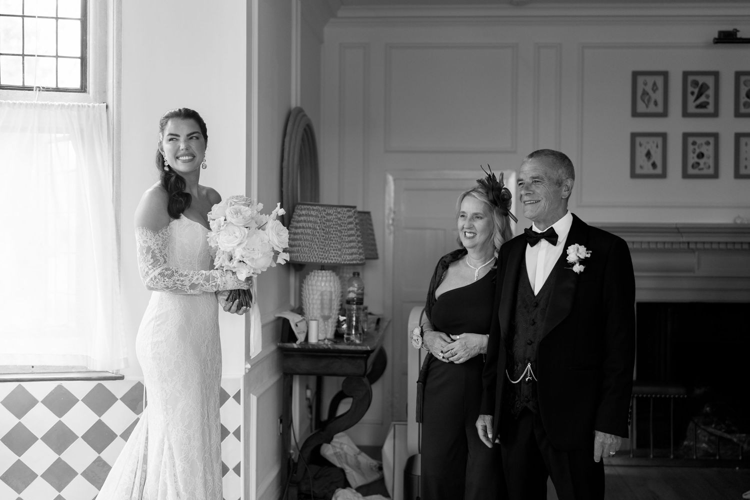 Black and white photo of a bride in lace gown standing with formally dressed guests in an elegant room.