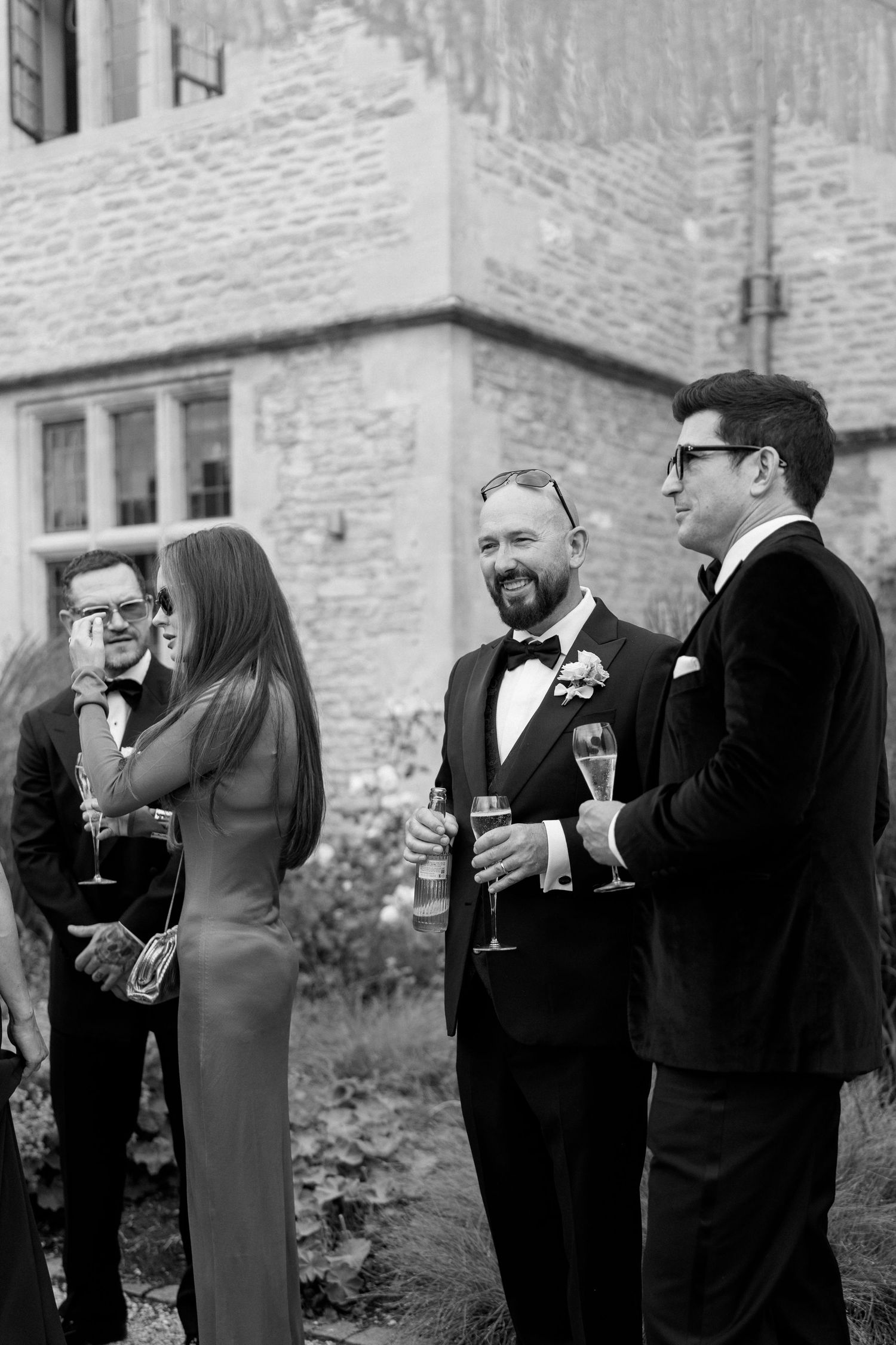 Black and white photo of wedding guests in formal attire socializing with champagne outside a stone venue.