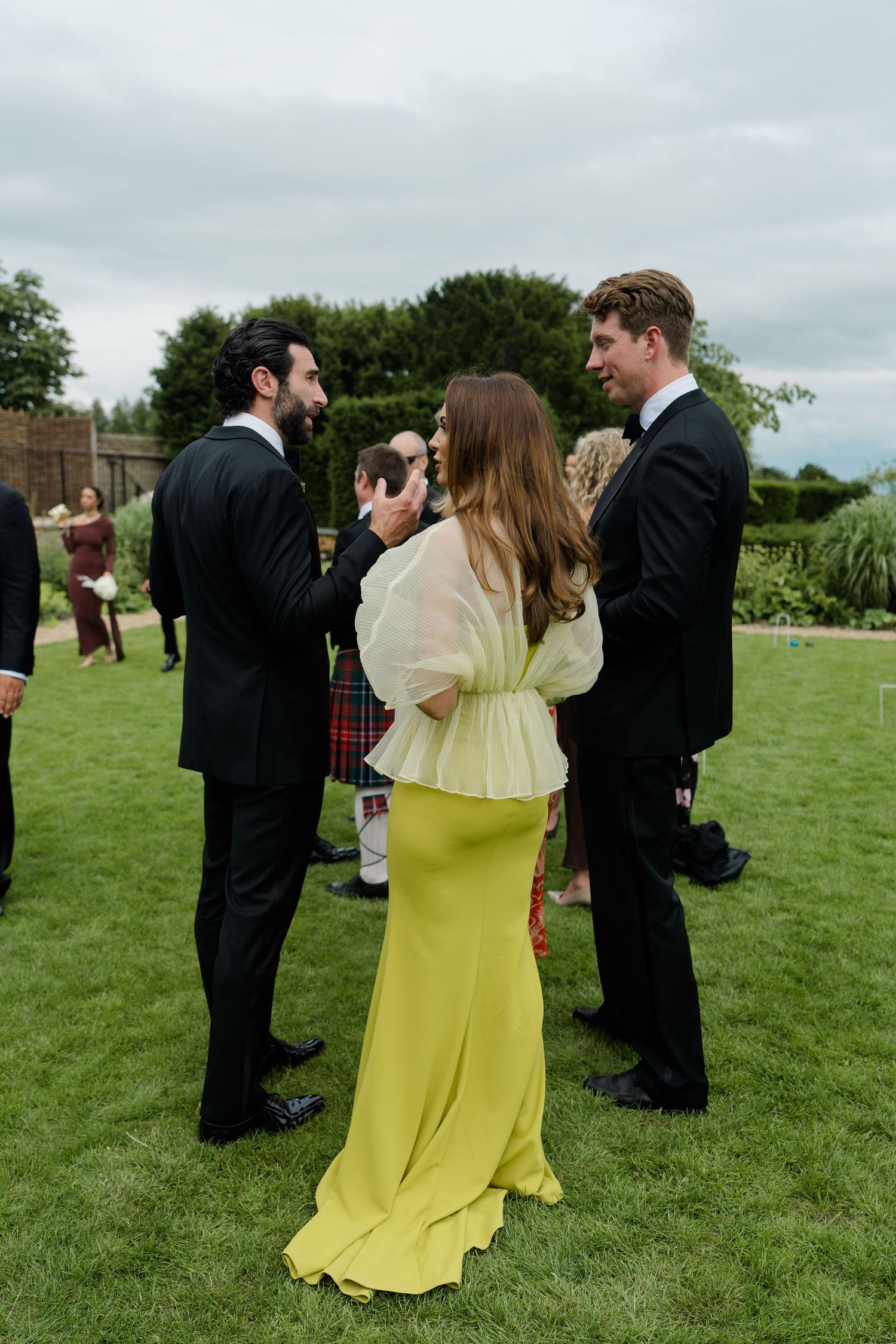 Wedding guests mingling on green lawn, woman in yellow dress conversing with two men in black tuxedos.