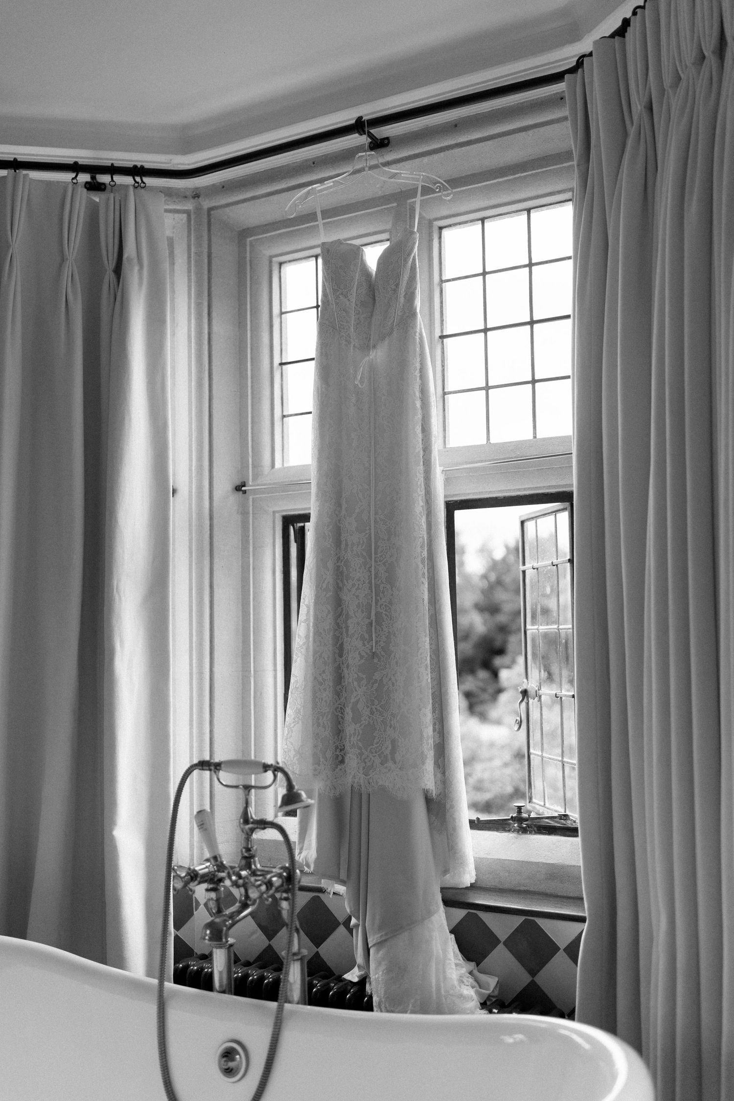 Elegant lace wedding dress hanging in window above freestanding bathtub, black and white photo.