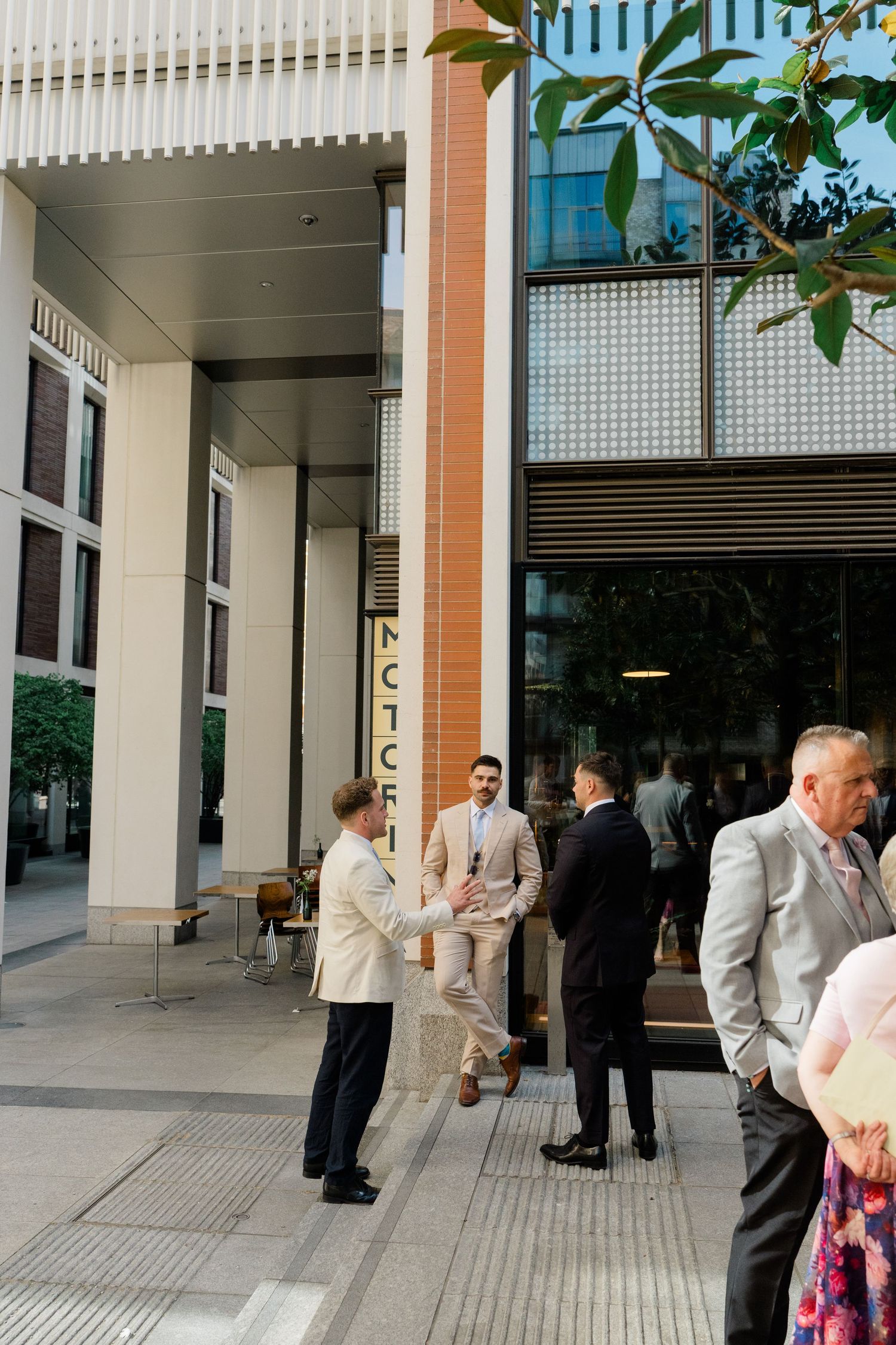 Elegantly dressed wedding guests in suits socializing outside a modern building on a sunny day.